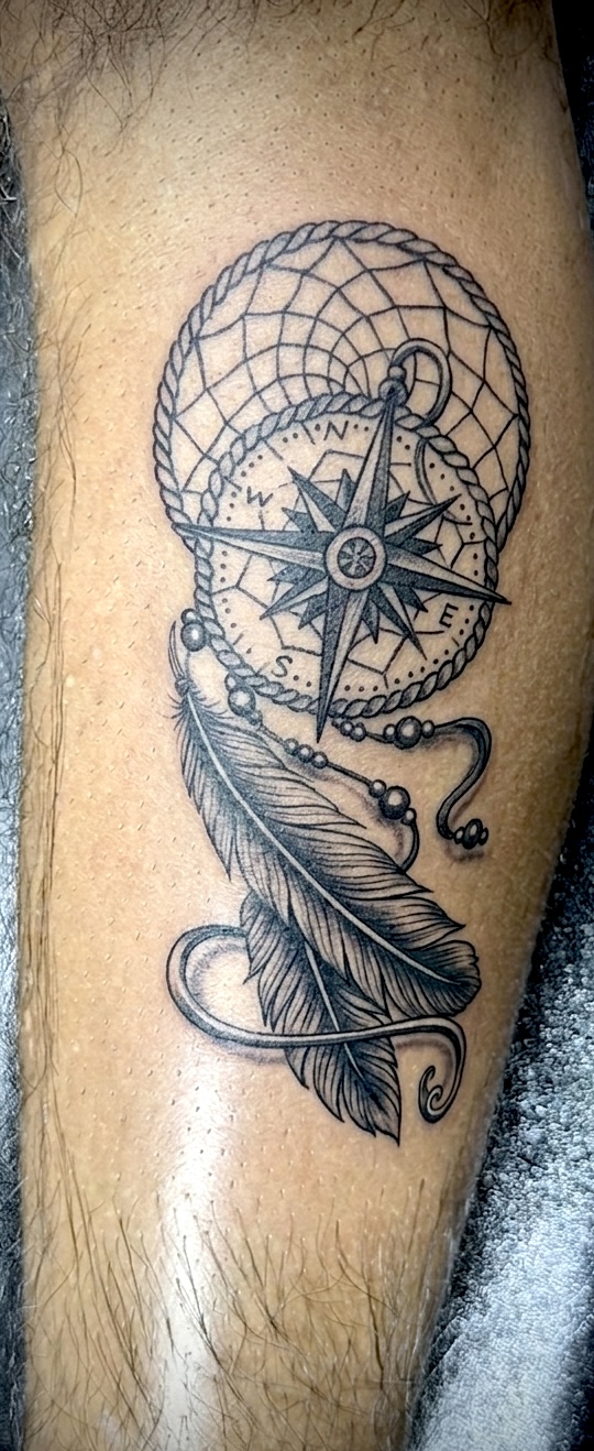 Tattoo of a dreamcatcher with a compass, feather, and beads on skin.