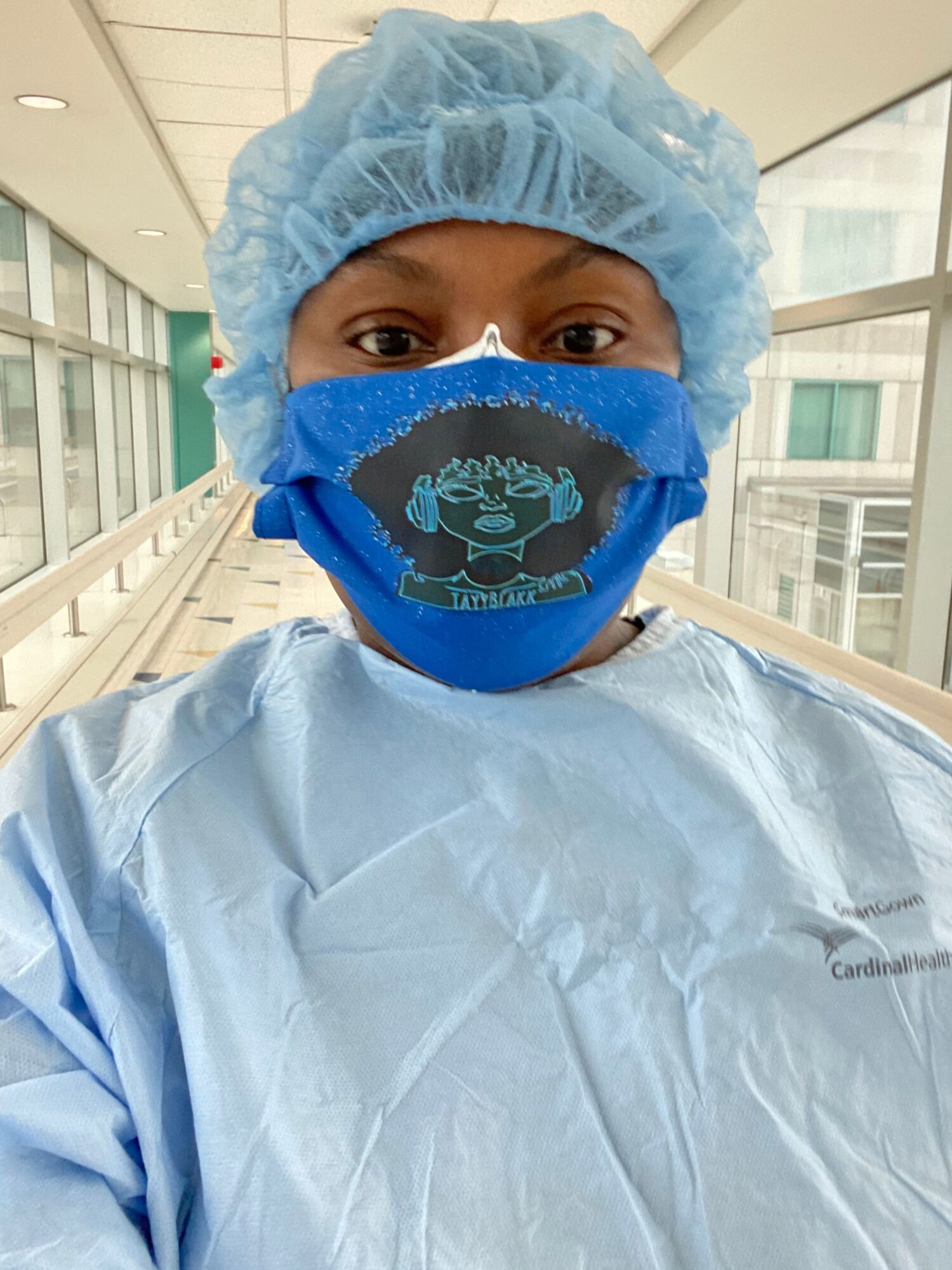 Person in medical attire wearing a blue mask and cap, standing in a hospital corridor.