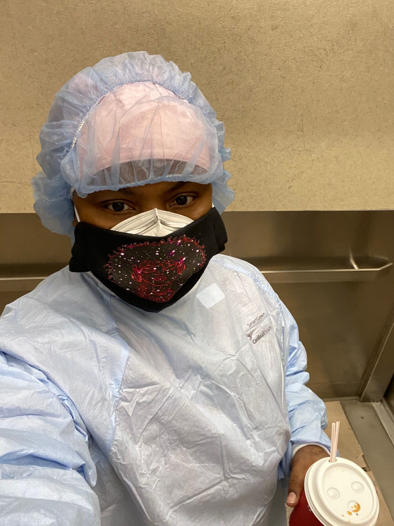 Person in medical protective clothing, mask, and hair cover holding a cup, lying on a hospital bed.