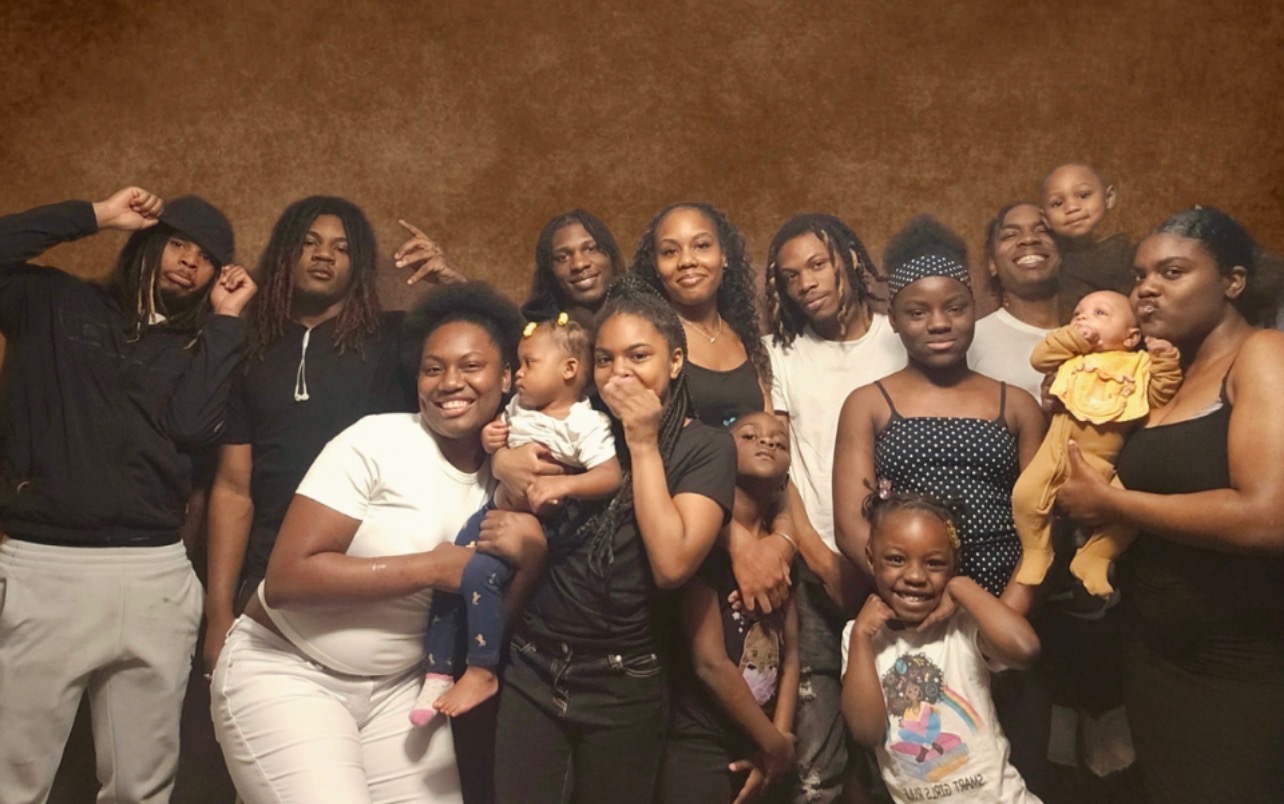 Group of people, including children, posing together indoors against a brown background, smiling and holding children.