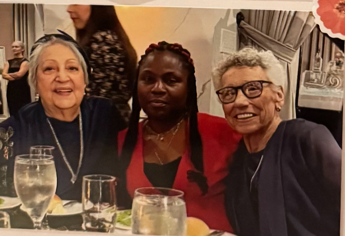 Three women sitting at a table, smiling, with drinks in front of them, in a social setting.