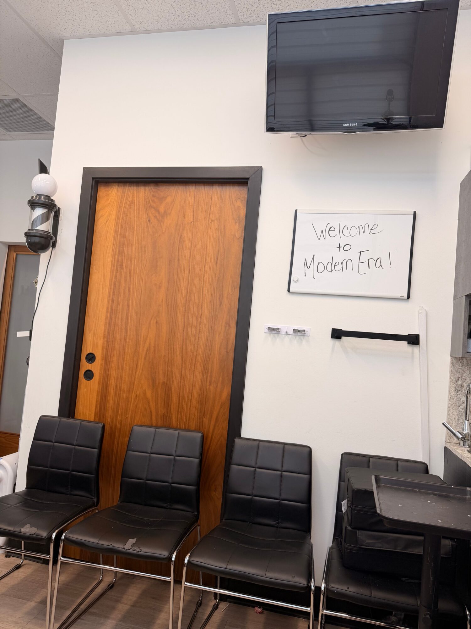 Waiting area with black chairs, a wooden door, and a whiteboard that says 'Welcome to Medical Office'.