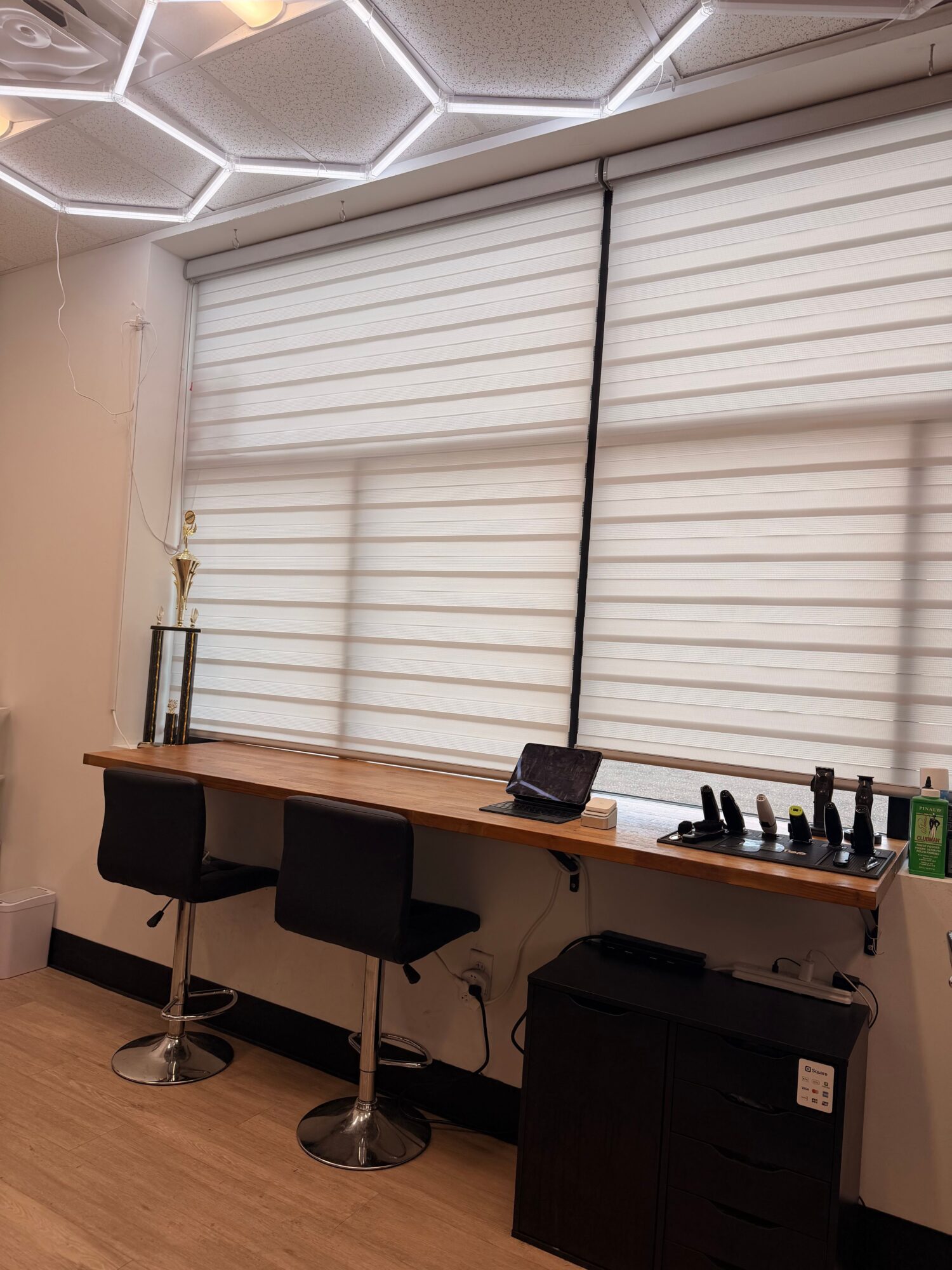 Office space with a desk, computer, and chairs, viewed from the side with window blinds in the background.