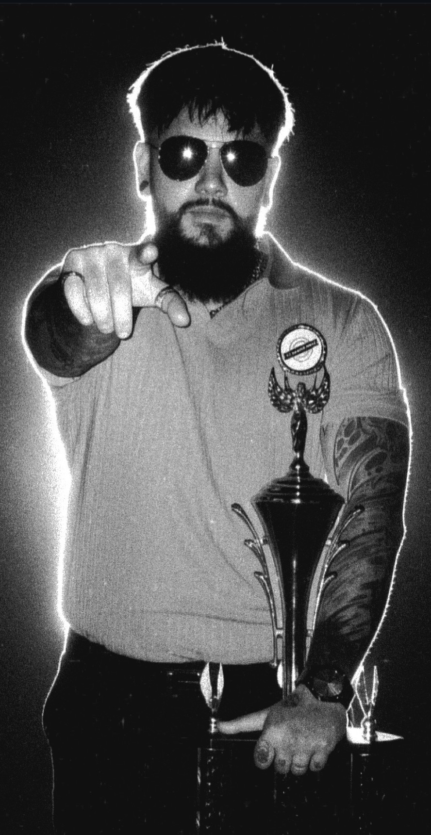 Man with sunglasses and beard pointing at camera, holding a trophy, with tattooed arm, in black and white.