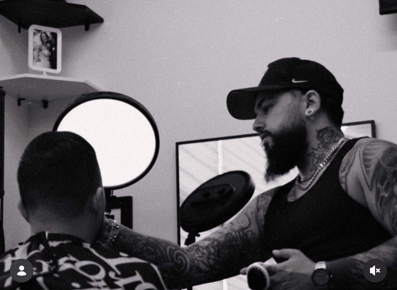 Man with tattoos and beard wearing a cap interacts with a child in a room with a computer monitor and shelves.