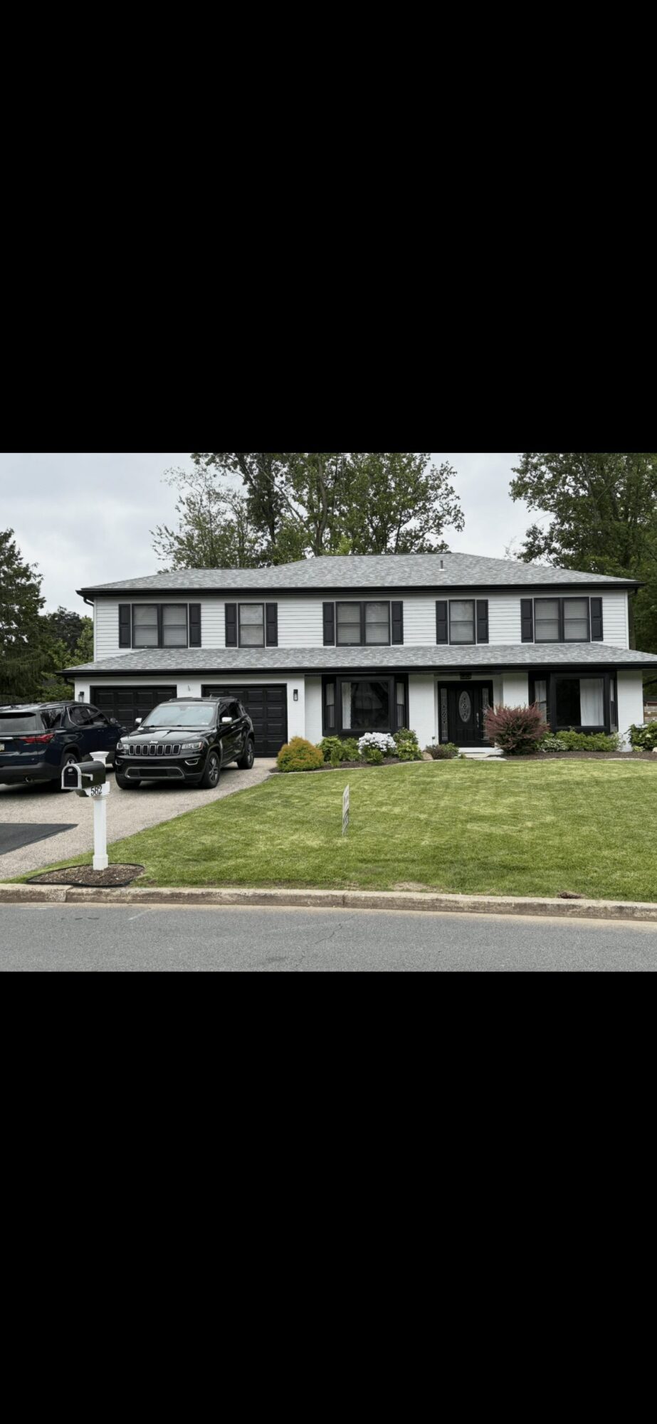 Two-story house with a front porch, surrounded by a lawn and trees, parked cars in driveway, cloudy sky.