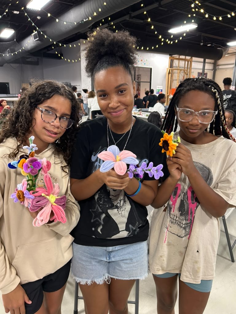 Three girls holding colorful craft flowers and a sunflower, standing indoors with string lights overhead.