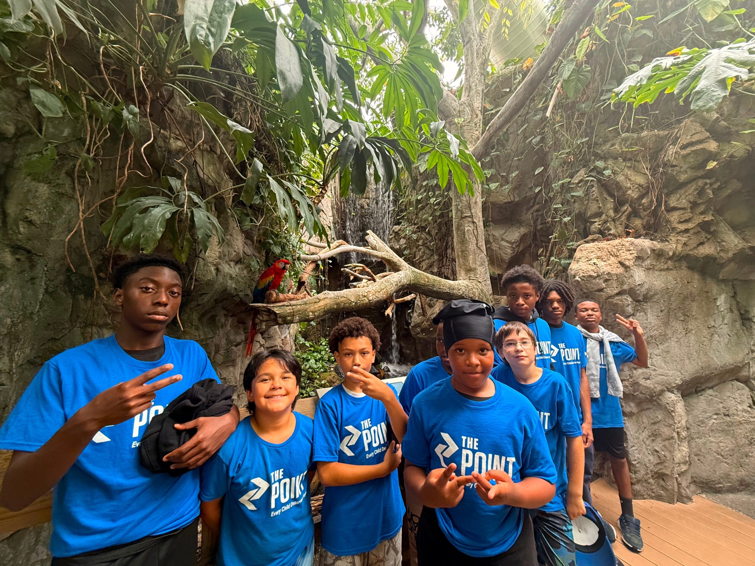 Group of children in blue shirts standing outdoors among rocks and trees, smiling and posing for the photo.