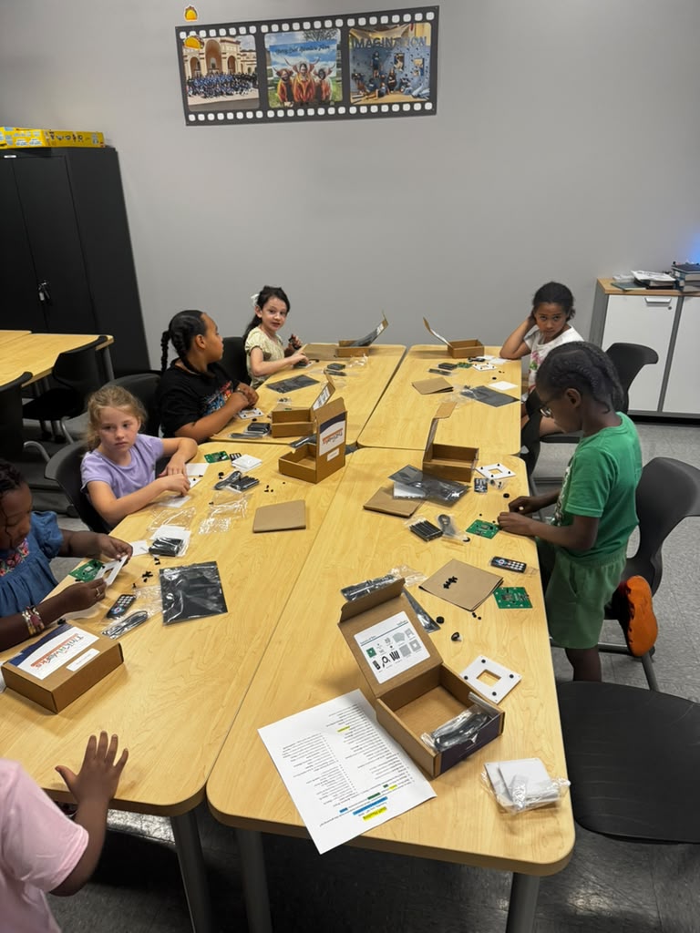 Children sit around a table with electronic devices and boxes, in a classroom with a gray wall and a poster, some children are smiling.