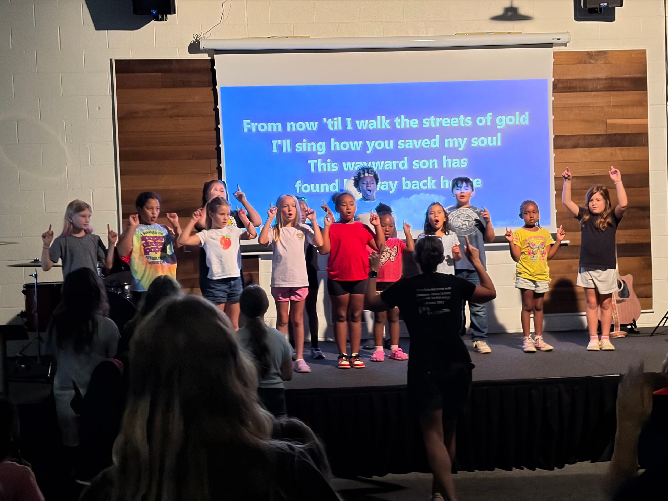 Group of children performing on stage with a blue screen displaying song lyrics behind them, audience watching.