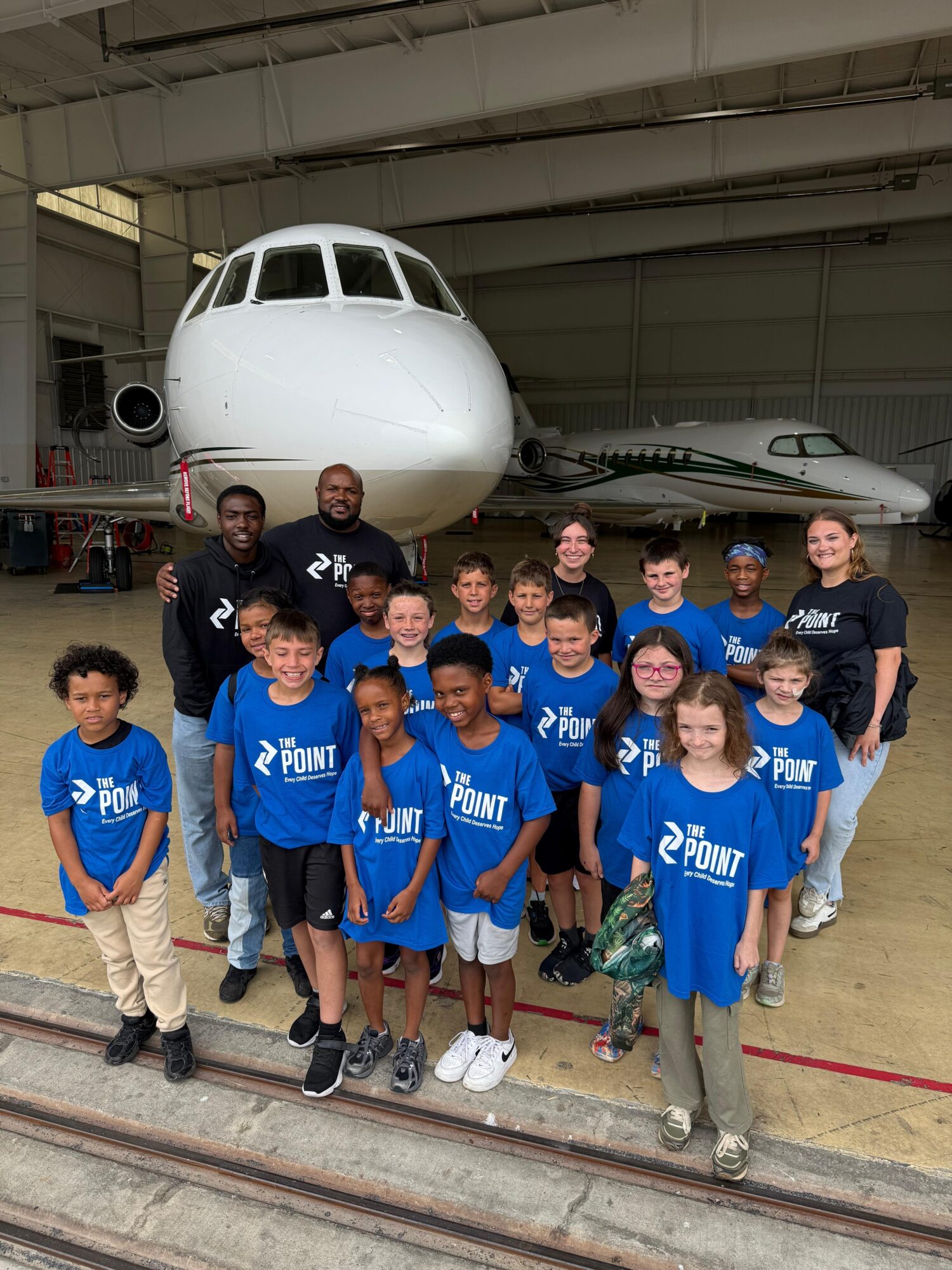 Group of children and adults in blue shirts posing in front of an airplane inside a hangar.