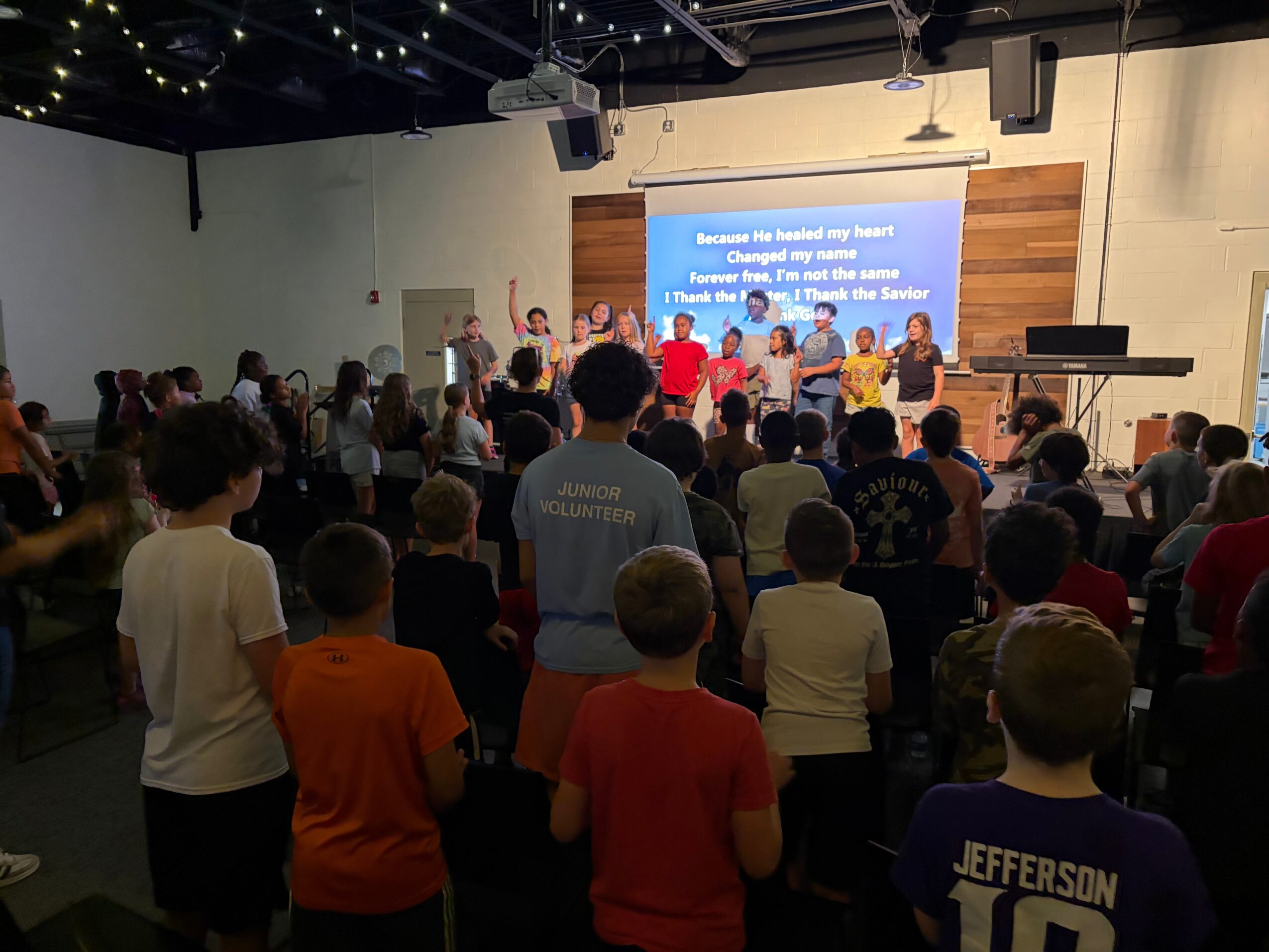 Crowd of people facing a stage with a large screen displaying text, in an indoor auditorium or church setting.