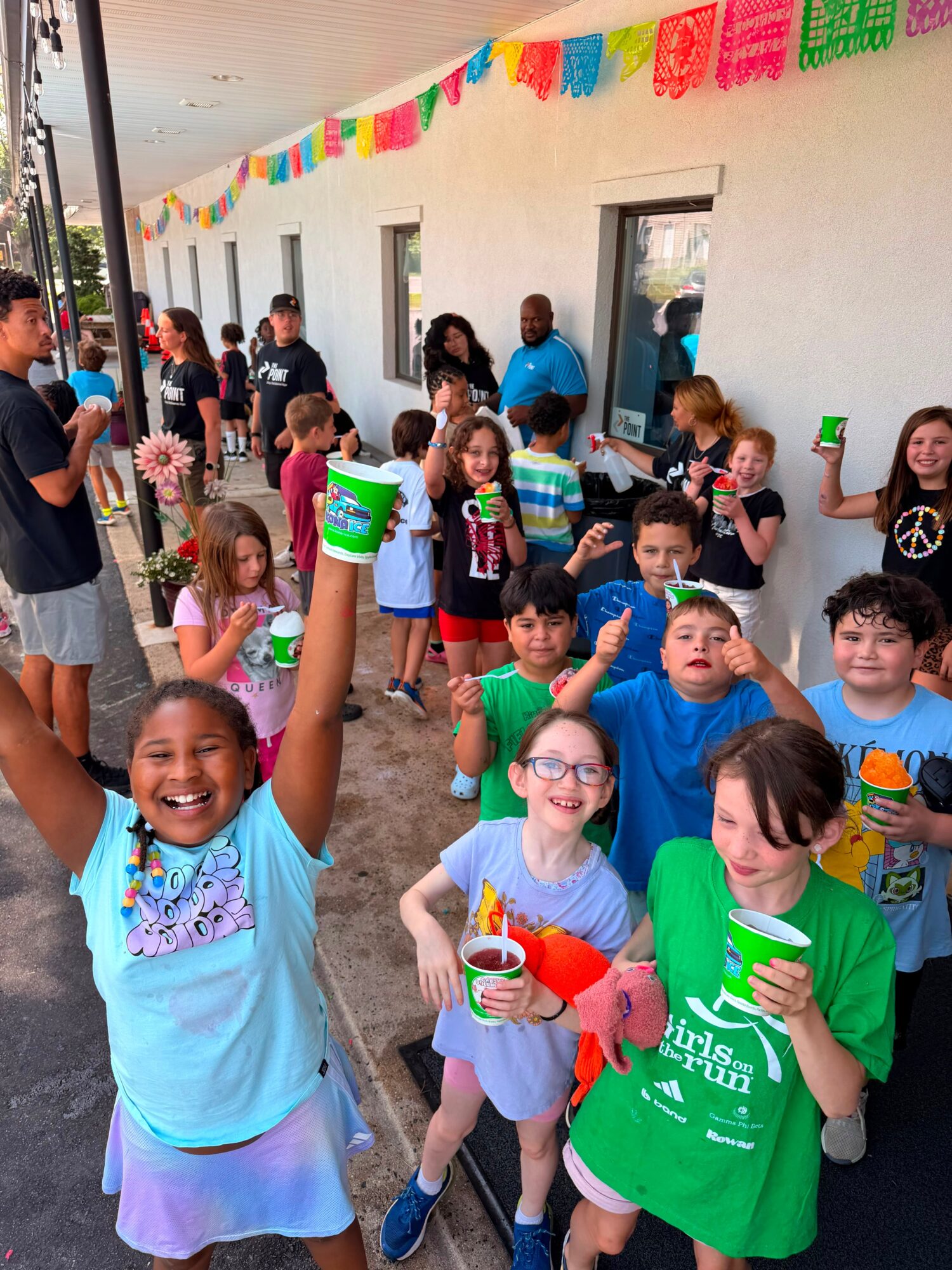 Children and adults gather outdoors under colorful decorations, holding drinks and smiling at a party.