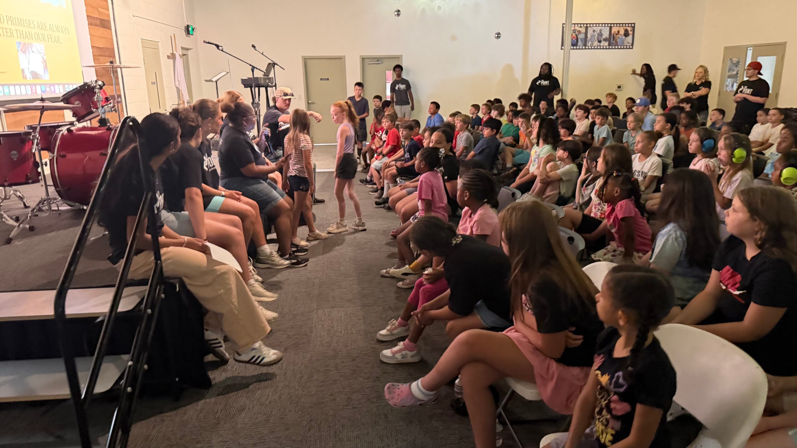 Large group of children and adults seated in a room, watching a performance or presentation at the front.