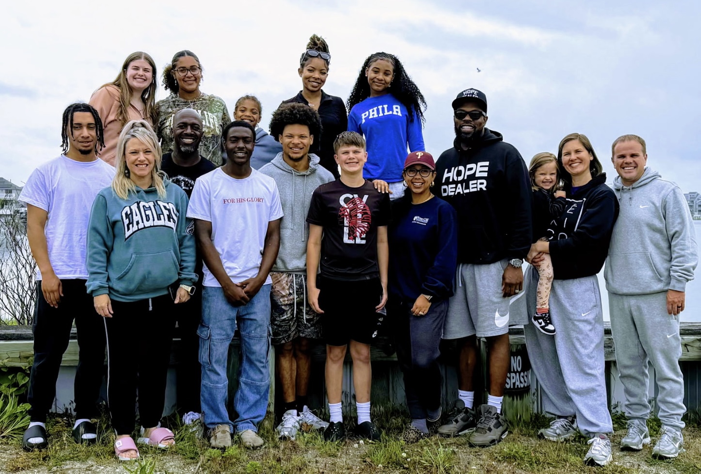Group of diverse young people outdoors, standing on grass with trees and sky in background.
