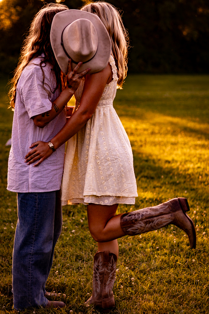 Two people embrace outdoors, one with a hat covering their face, the other in a light dress, during sunset.