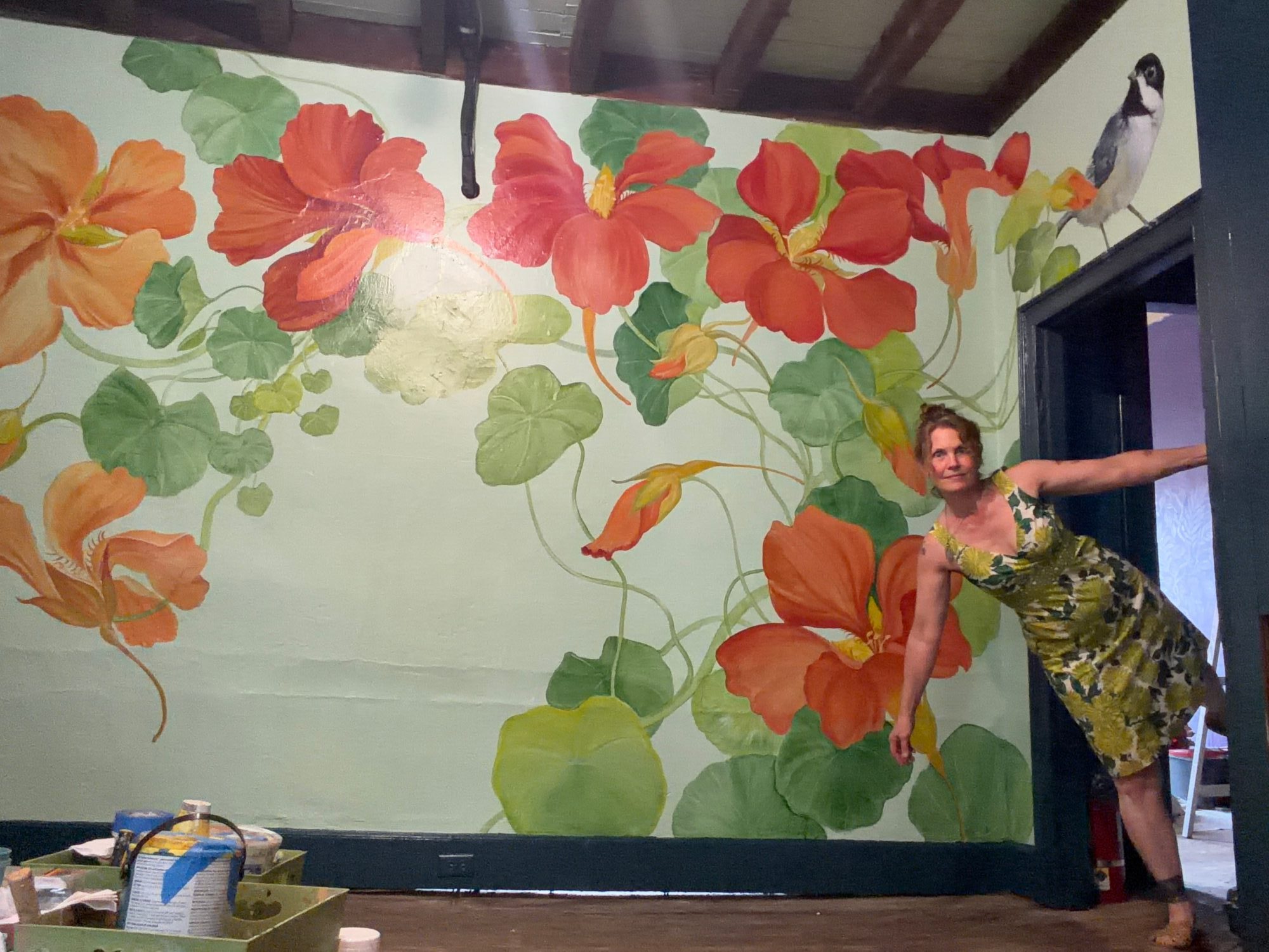 Person standing next to a large floral mural with orange and red flowers and green leaves on a wall.