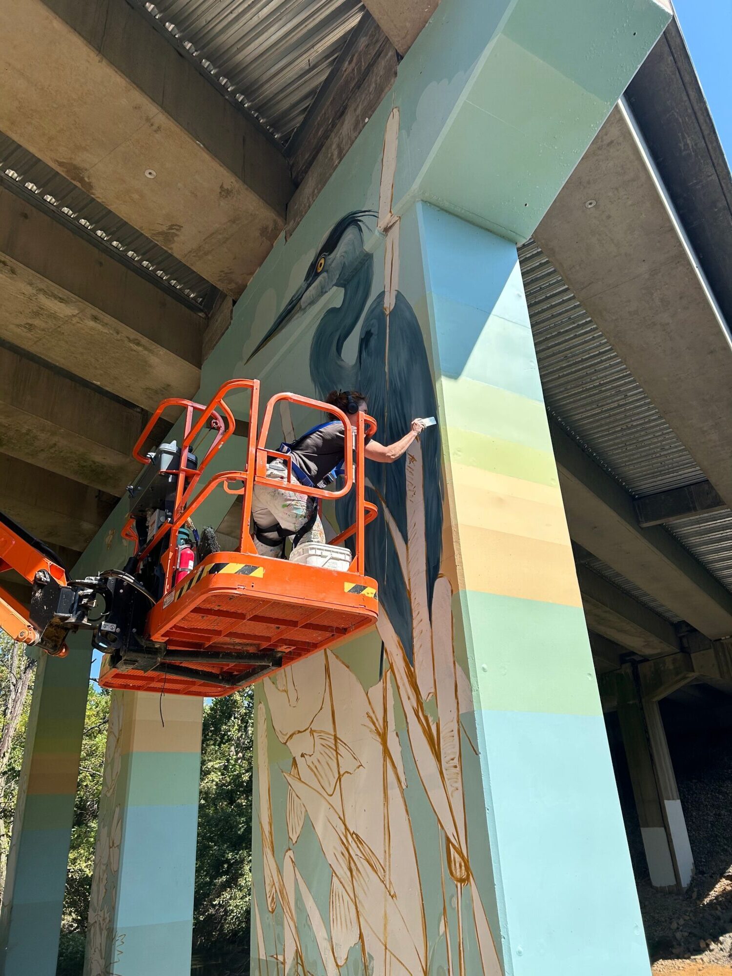Person on an orange lift working on a large mural with a colorful abstract design, outdoors, with a building in the background.