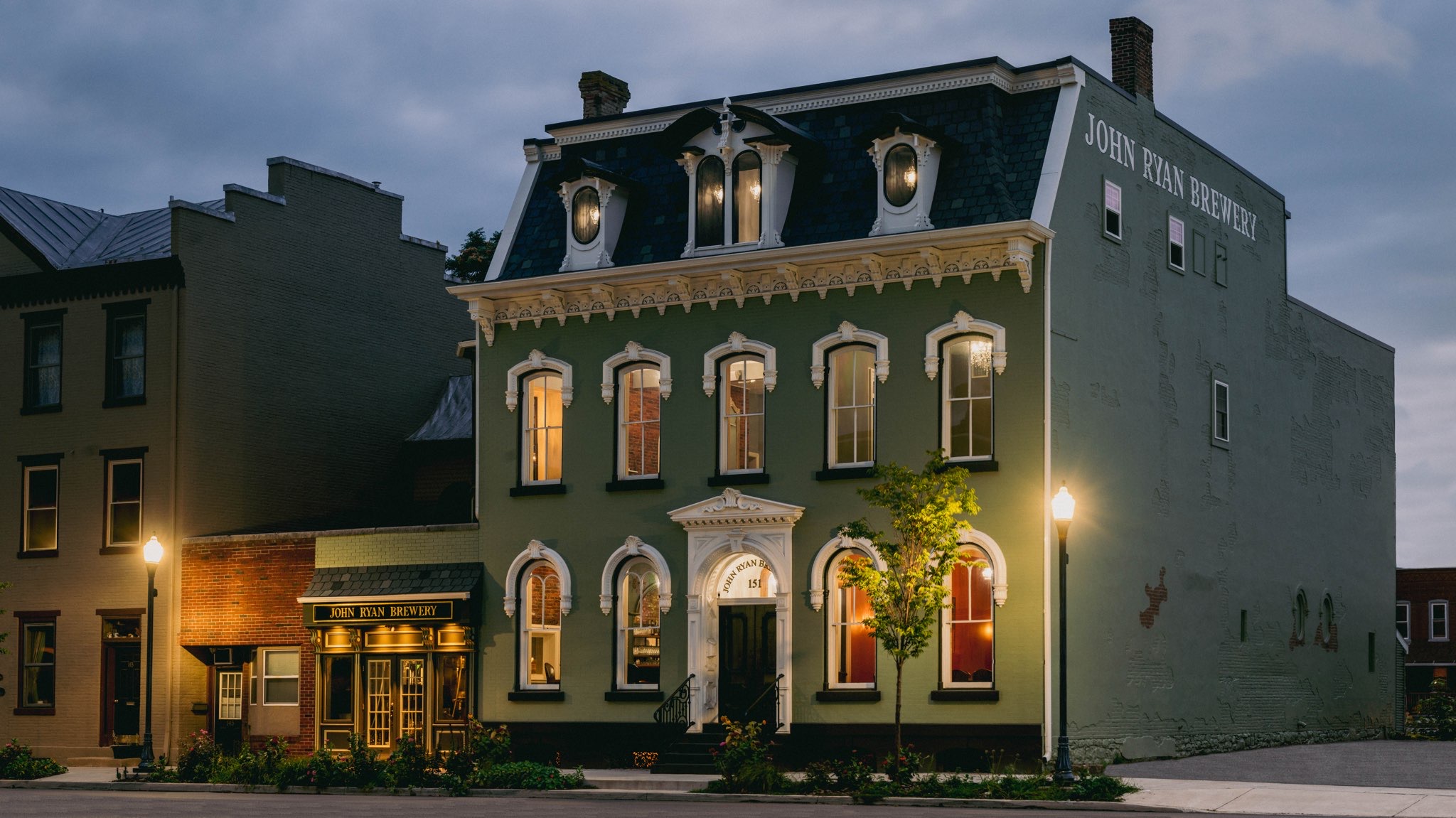 Three-story building with ornate windows, a small tree, and streetlamp in front, at dusk.