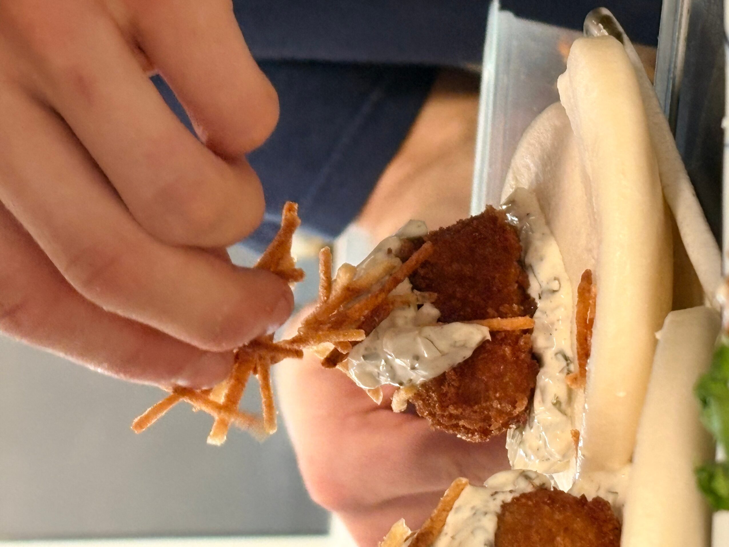 Person holding a sandwich with fried chicken, lettuce, and sauce, with a hand reaching for the food.
