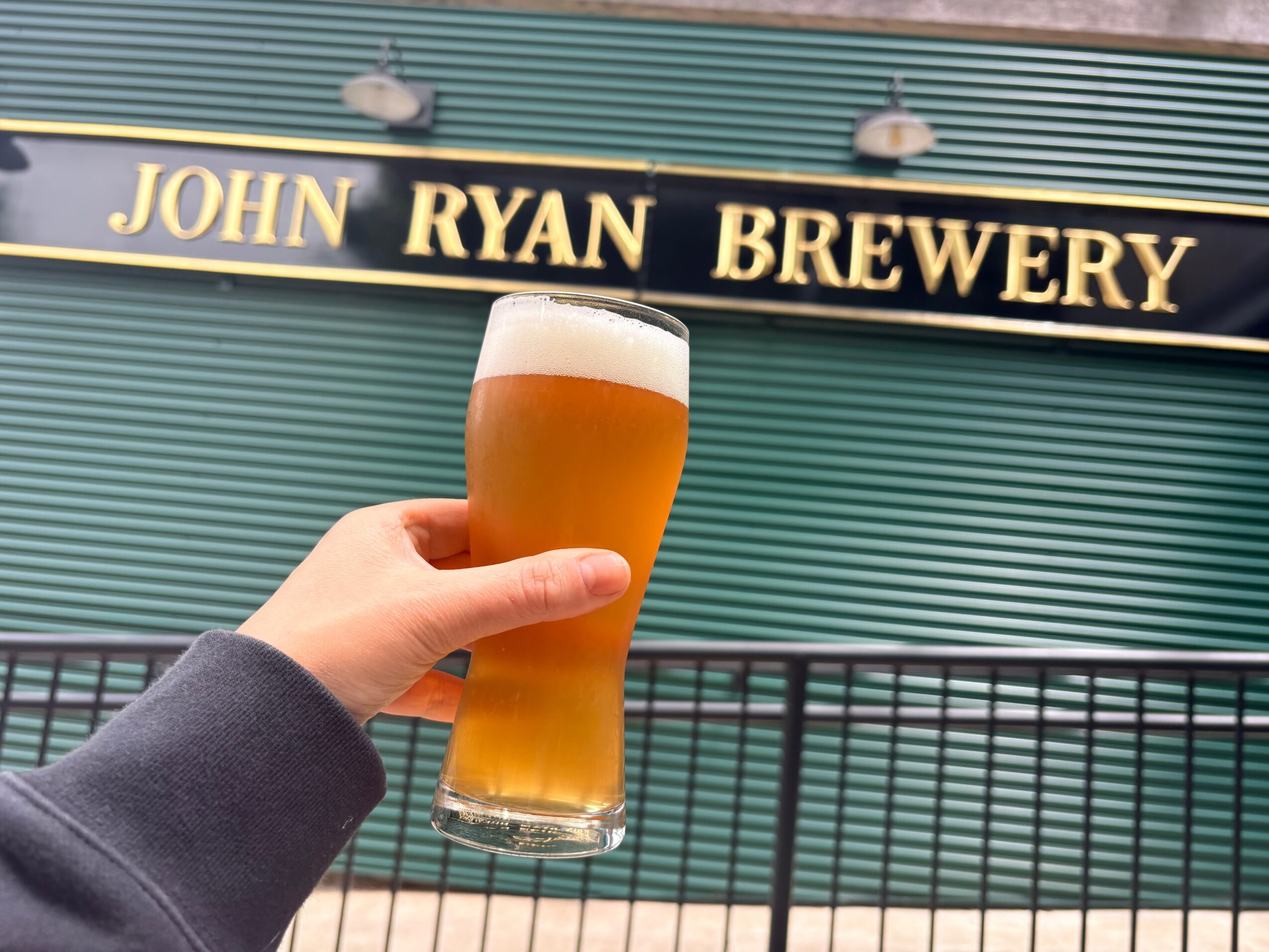 Hand holding a glass of beer in front of a brewery sign reading 'John Ryan Brewery'.