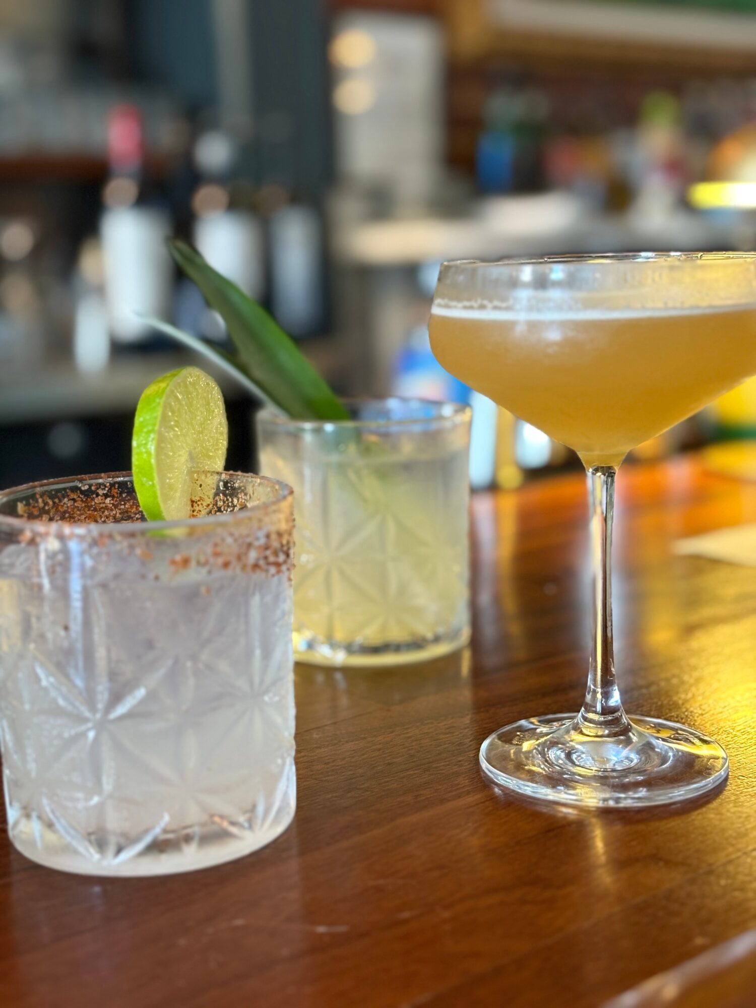 Three cocktails on a bar counter with blurred background, one garnished with lime, in various glass styles.