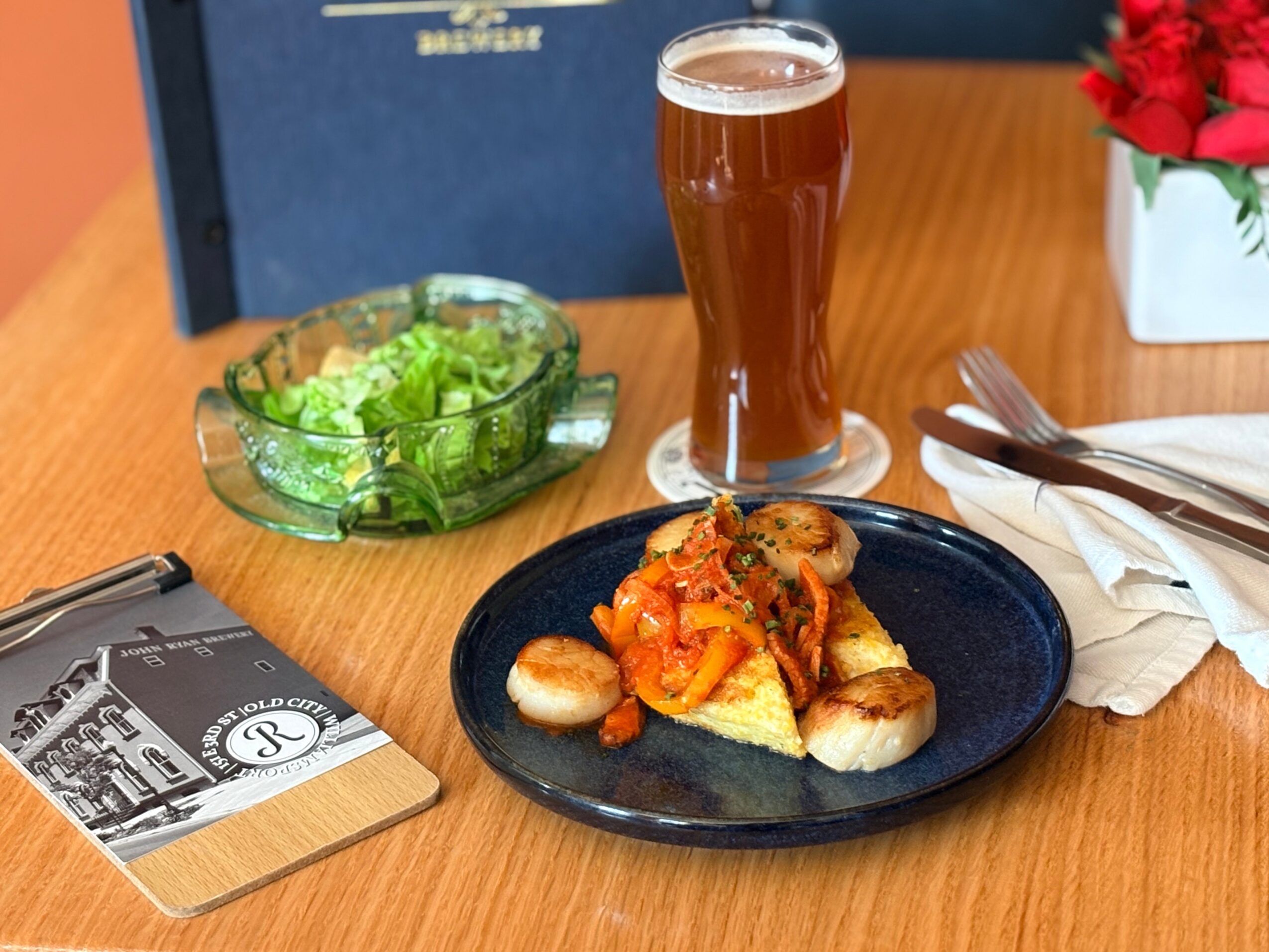 Plate with seafood and vegetables, glass of beer, salad bowl, napkin, and utensils on a wooden table.