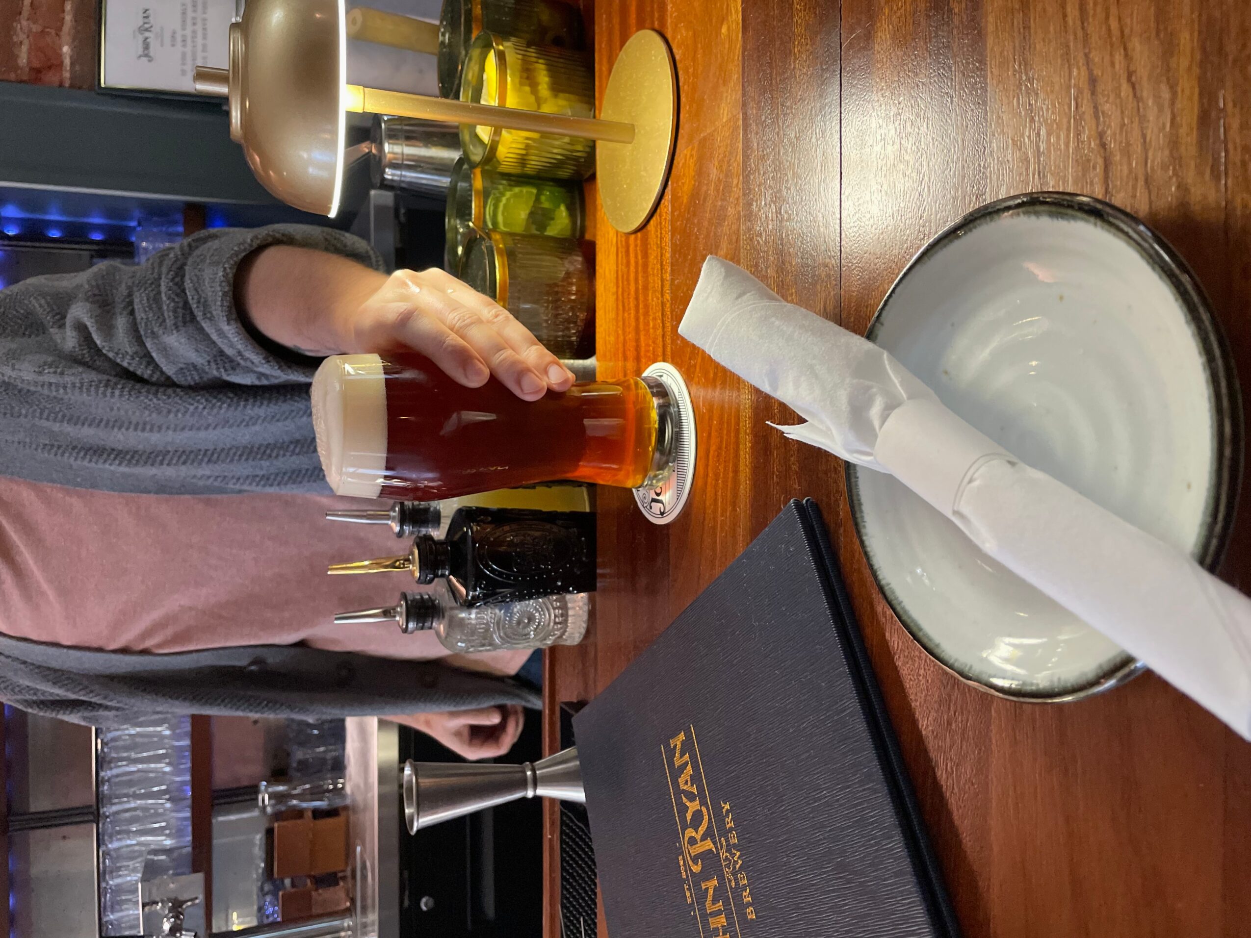 Person pouring beer into a glass at a bar counter with a napkin in a dish, various bottles and glasses in background.