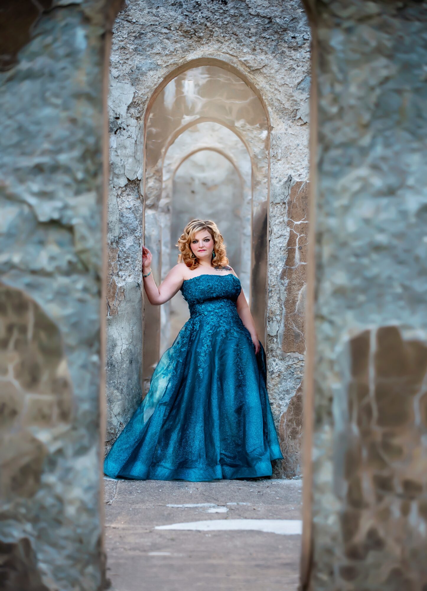 Woman in a blue gown standing in an arched stone doorway, holding the door frame, with stone walls around her.