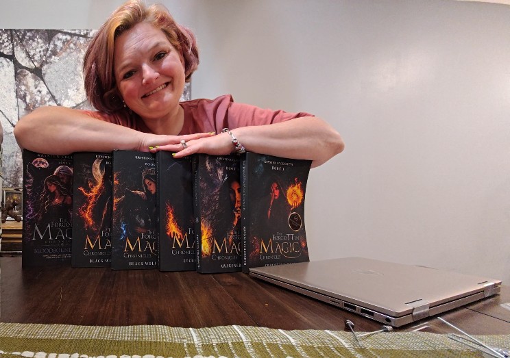 Woman with short hair smiling, resting her arms on five books with fiery covers, on a table with a laptop nearby.
