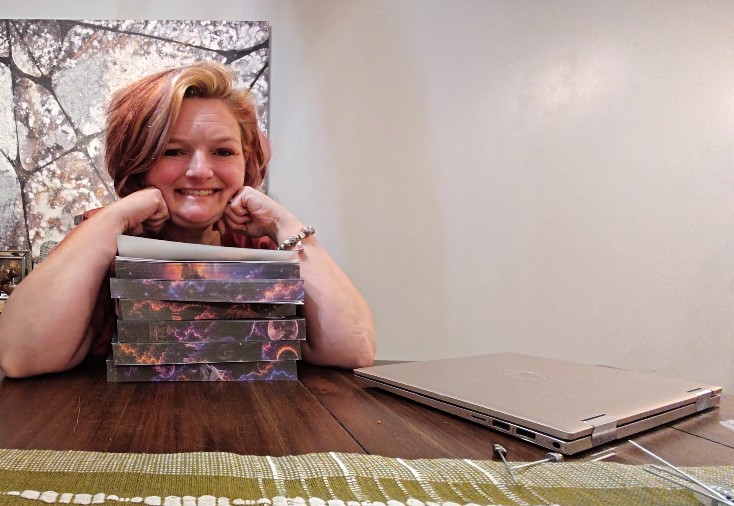 Smiling woman with red hair resting her chin on her hands, sitting at a table with a stack of books and a laptop.