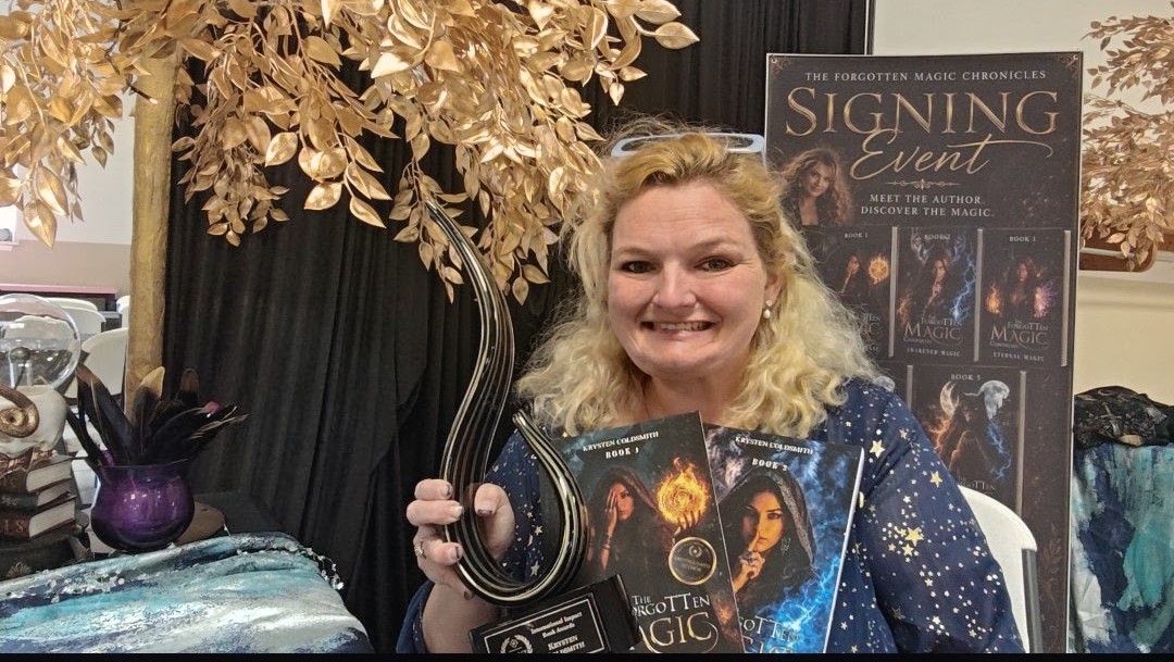Smiling woman holding a book at a signing event, with a poster and decorative plants in the background.