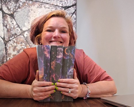 Woman smiling, holding a colorful book, sitting at a table indoors with a window showing trees outside.