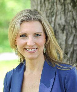 Woman with blonde hair smiling outdoors, wearing a blue jacket, standing near a tree.