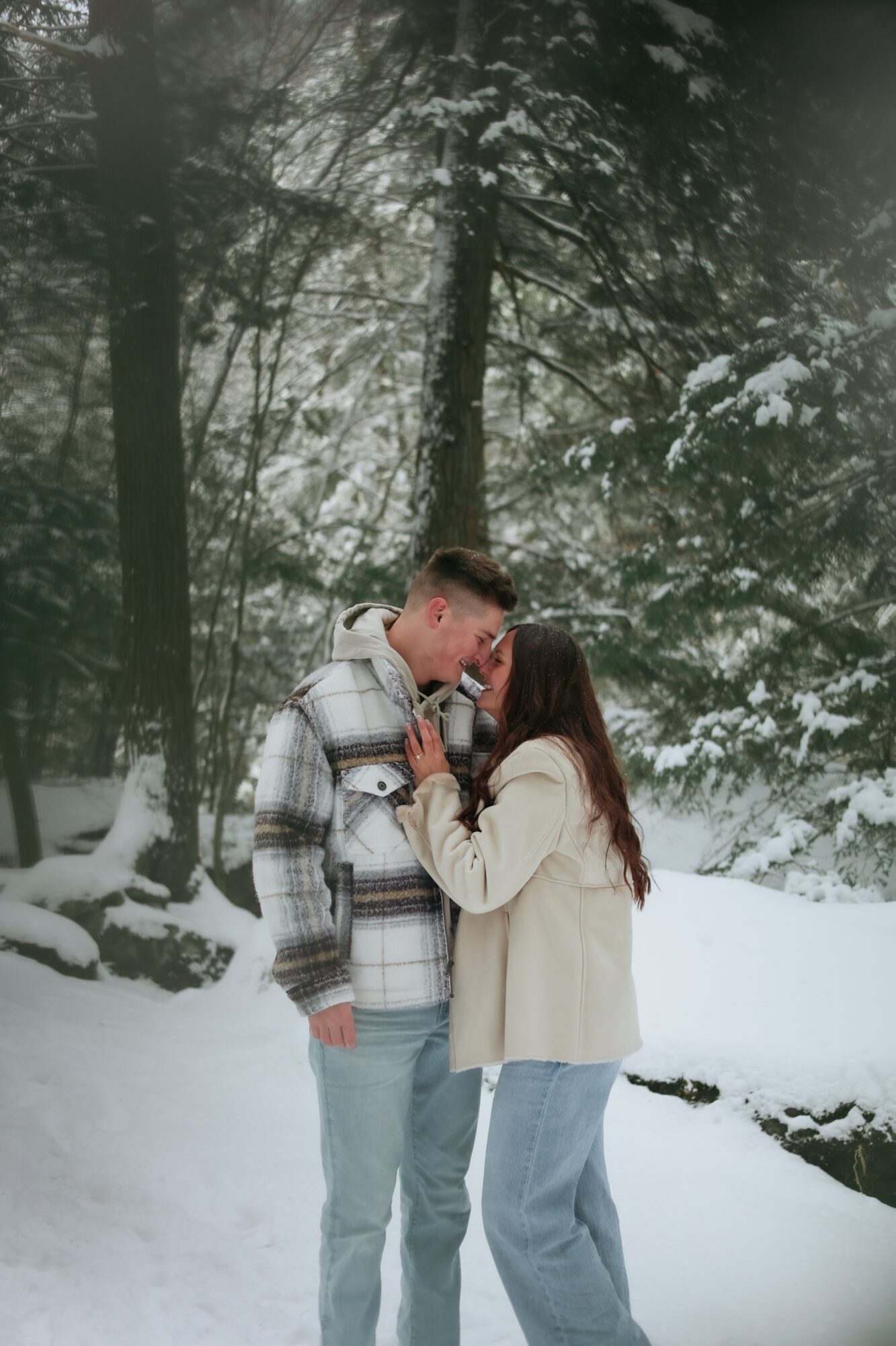 Two people standing close together in a snowy forest, touching foreheads and holding hands, surrounded by snow-covered trees.