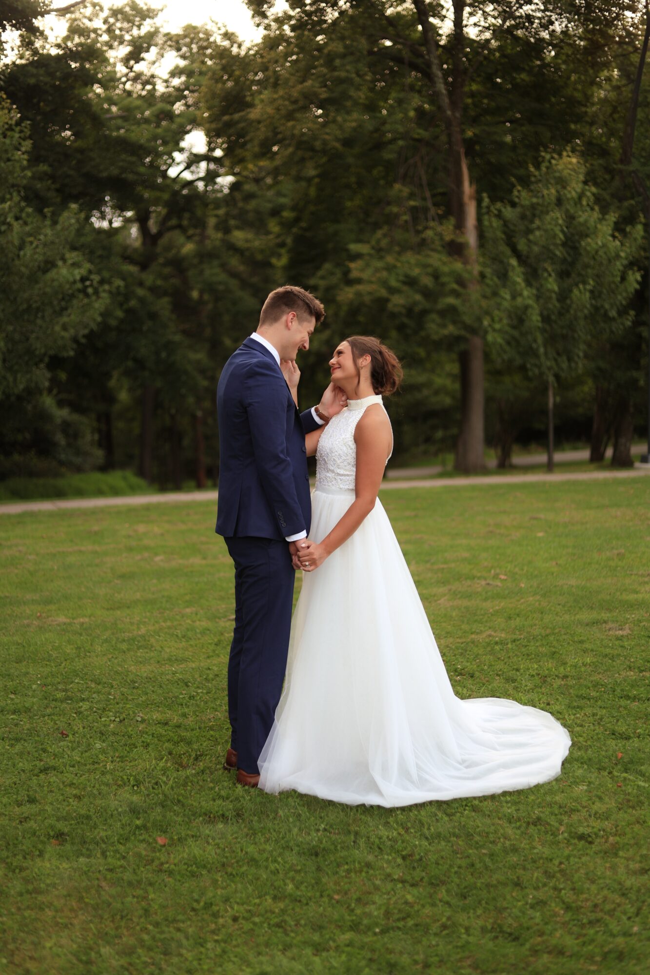 A bride and groom stand close together on a grassy field, holding hands and gazing at each other, with trees in the background.