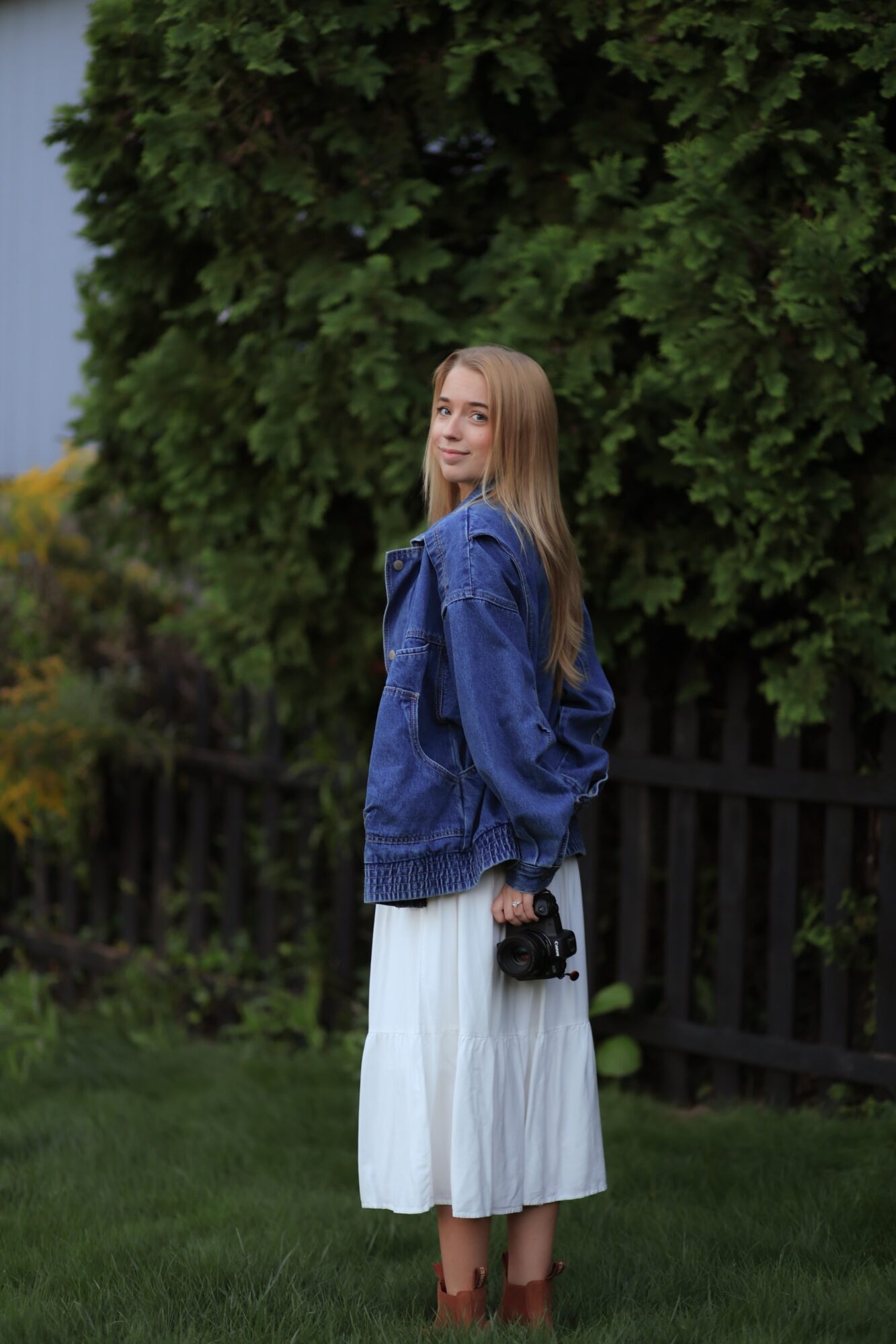Young woman with long hair standing outdoors near a black fence and large green trees, holding a camera, wearing a blue jacket and white skirt.