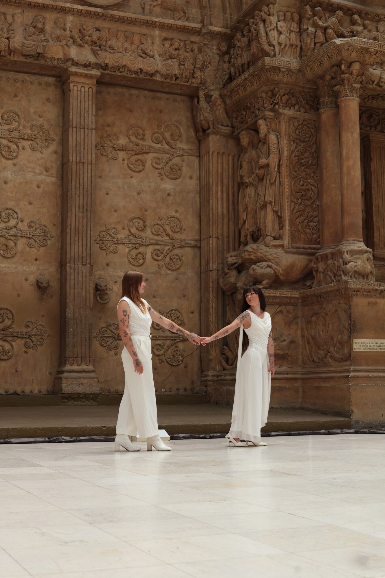 Two women in white dresses holding hands on stage with ornate, classical architecture background.