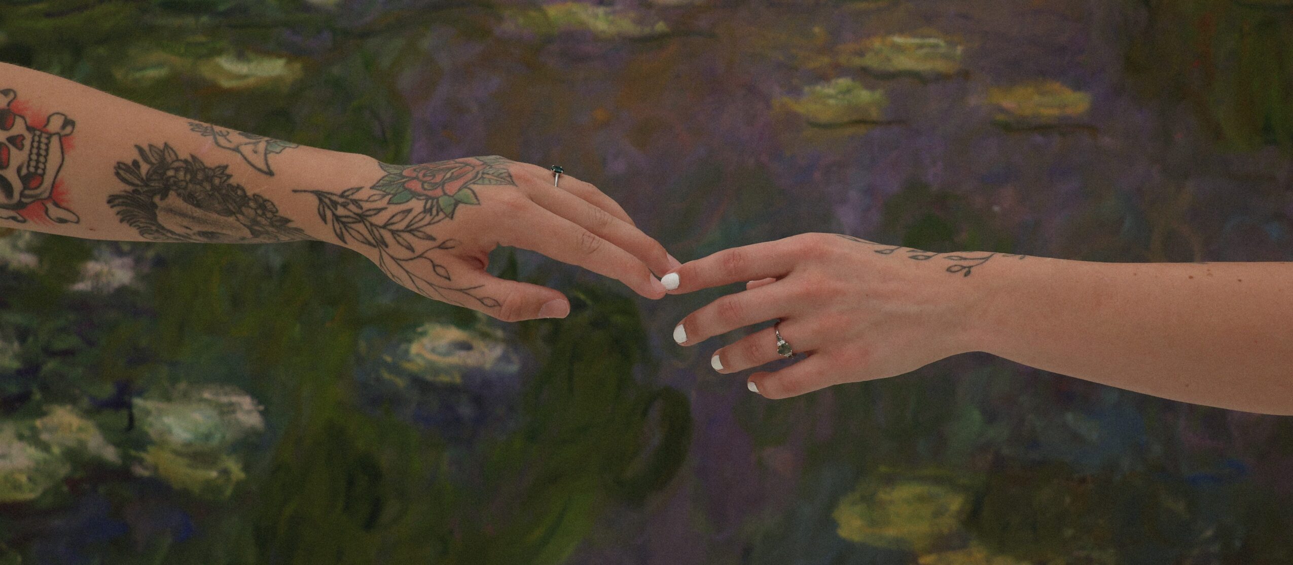 Two hands reaching towards each other over water, one with tattoos, the other with a ring, touching fingertips.