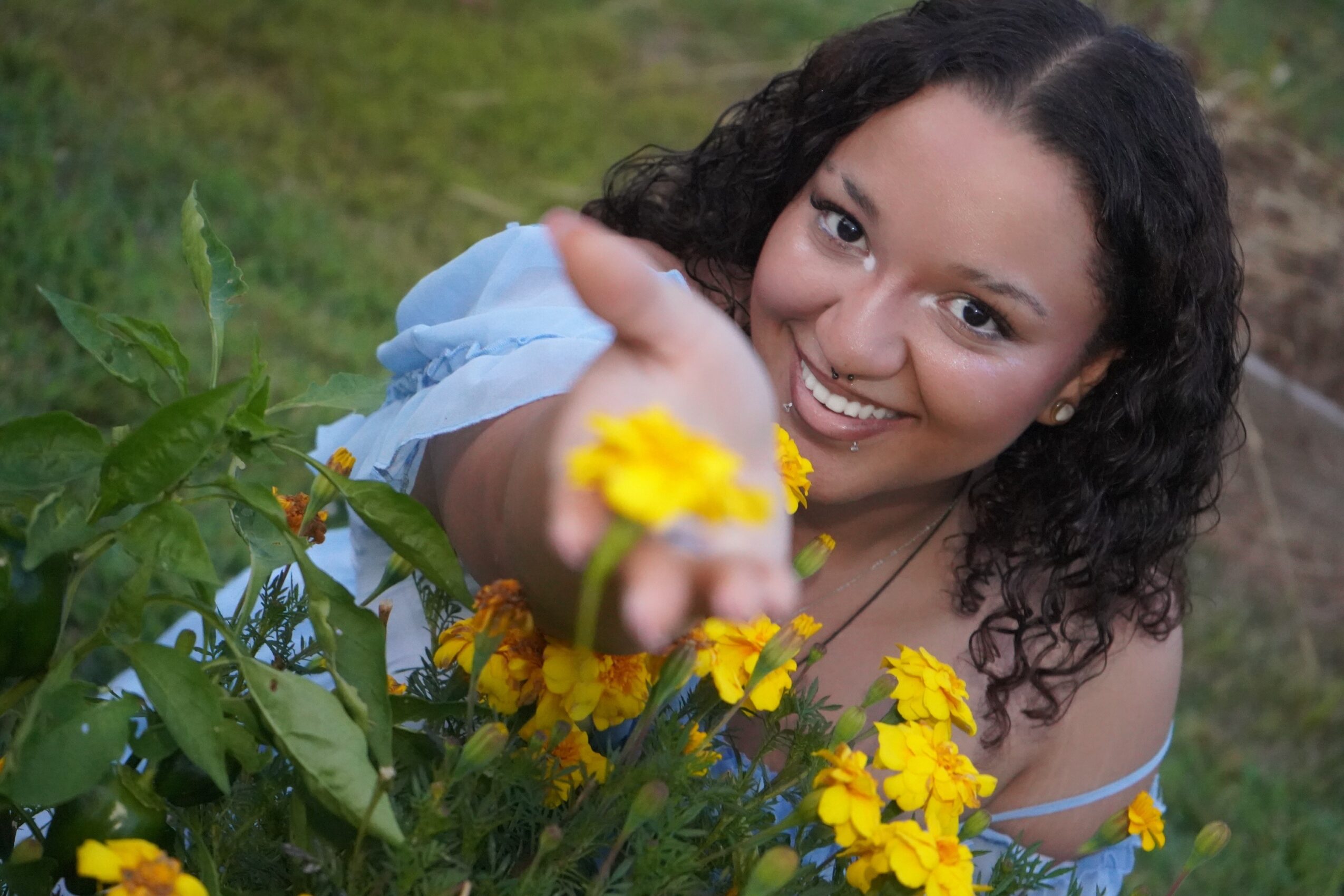 Smiling woman with curly hair reaching toward yellow flowers outdoors, surrounded by green plants.