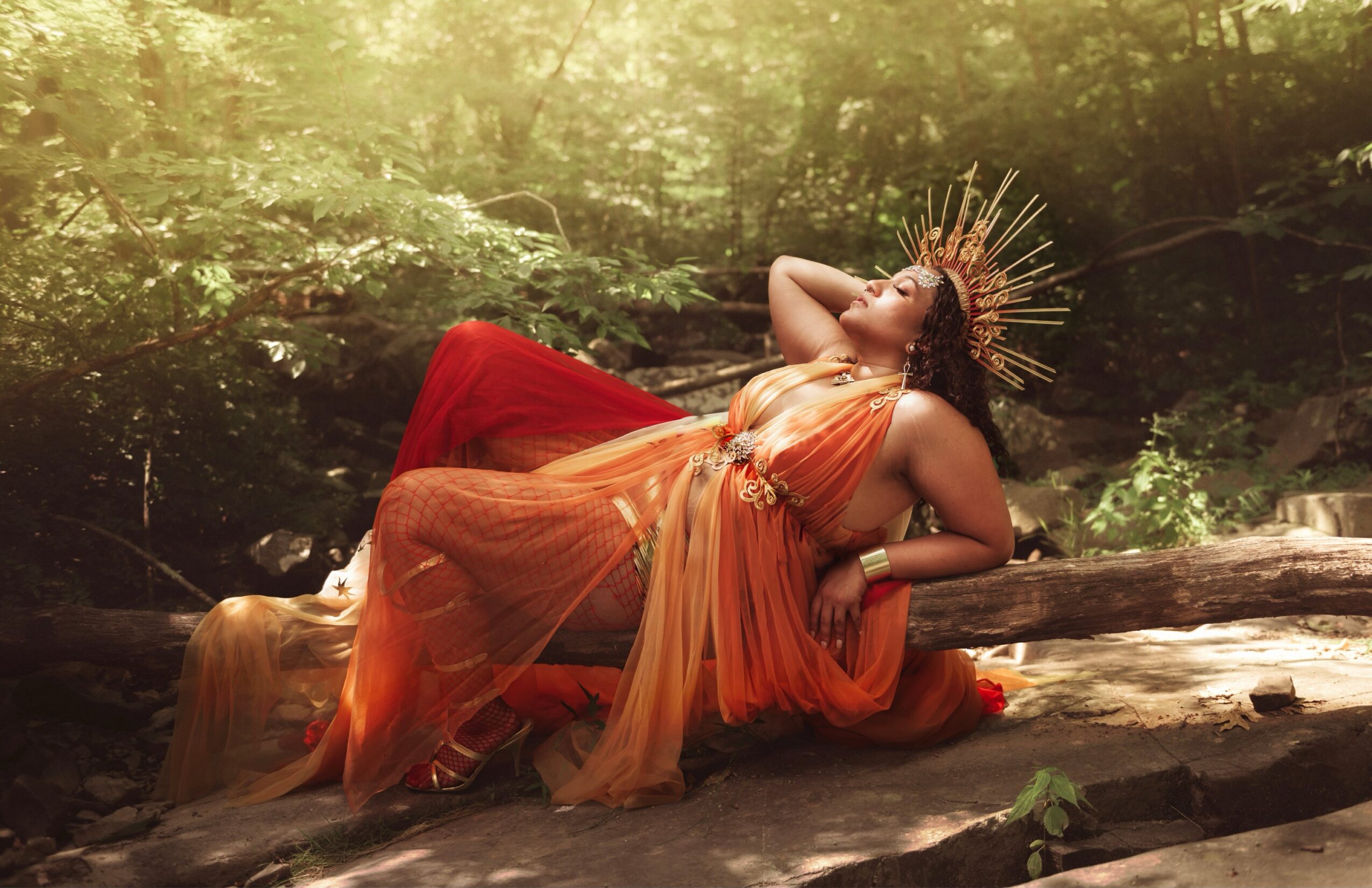 Woman in orange and red flowing dress reclining on a log in a forest, wearing a golden crown, sunlight filtering through trees.
