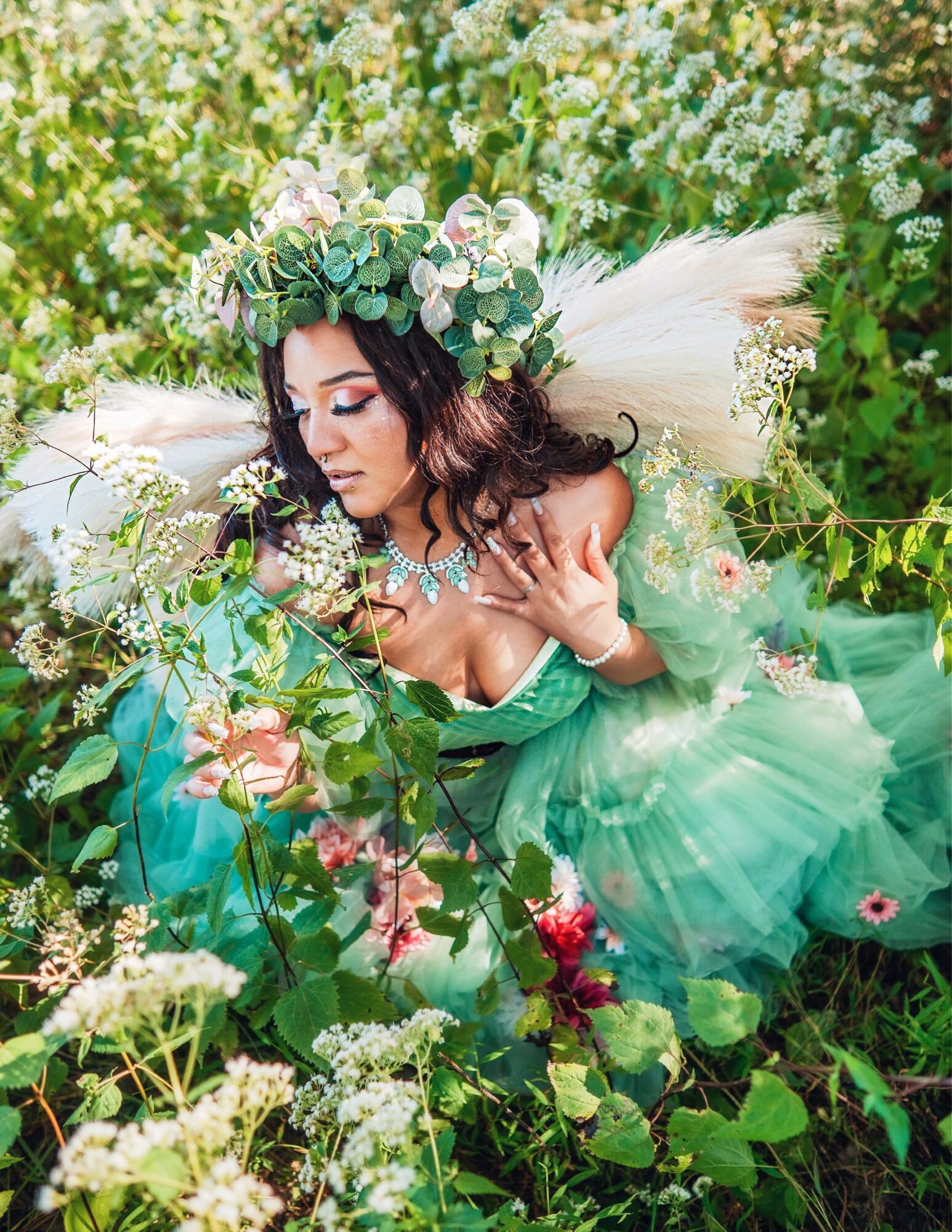 Woman in green dress with floral crown lying among white and pink flowers in a garden.
