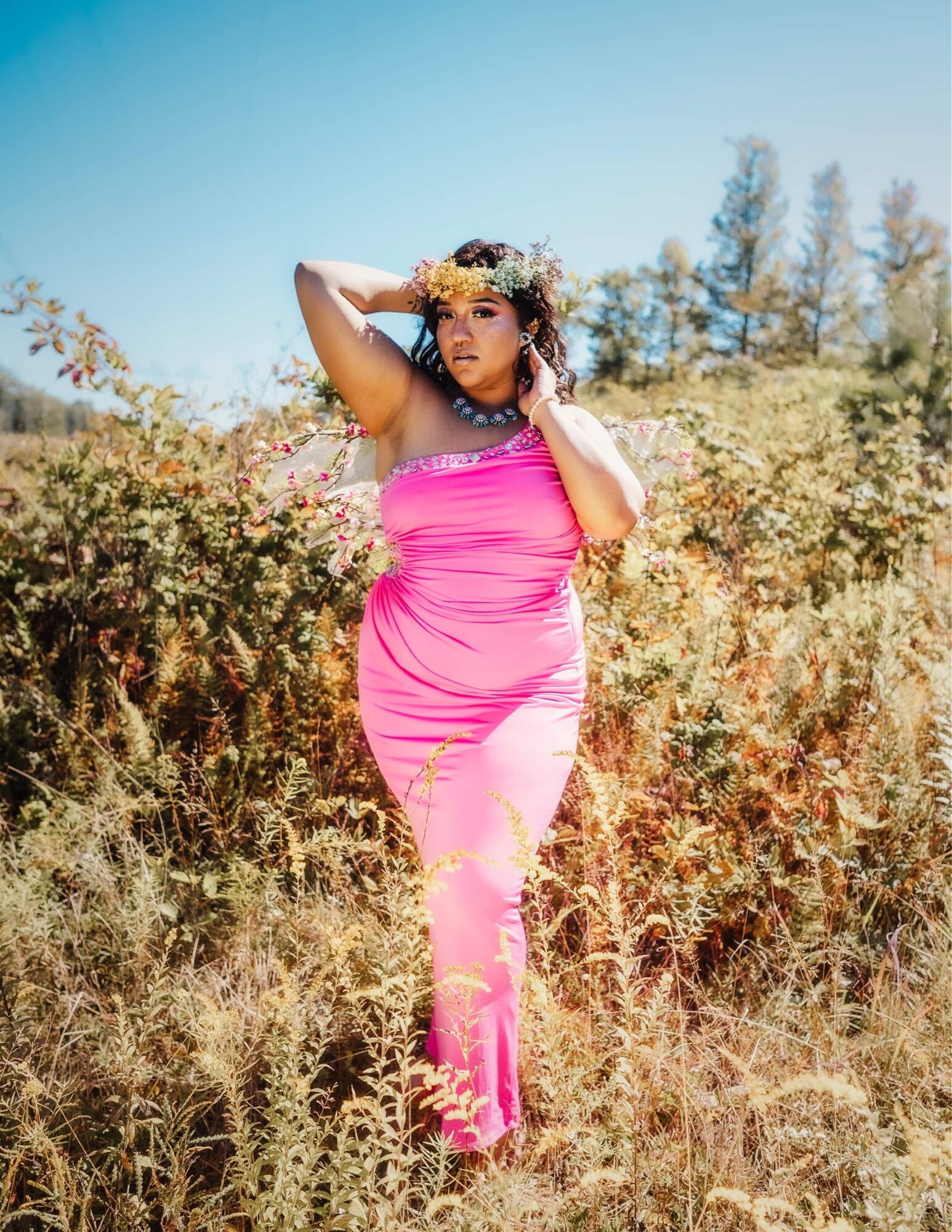 Woman in a pink dress standing outdoors in a field with trees and blue sky, holding her head with one hand.