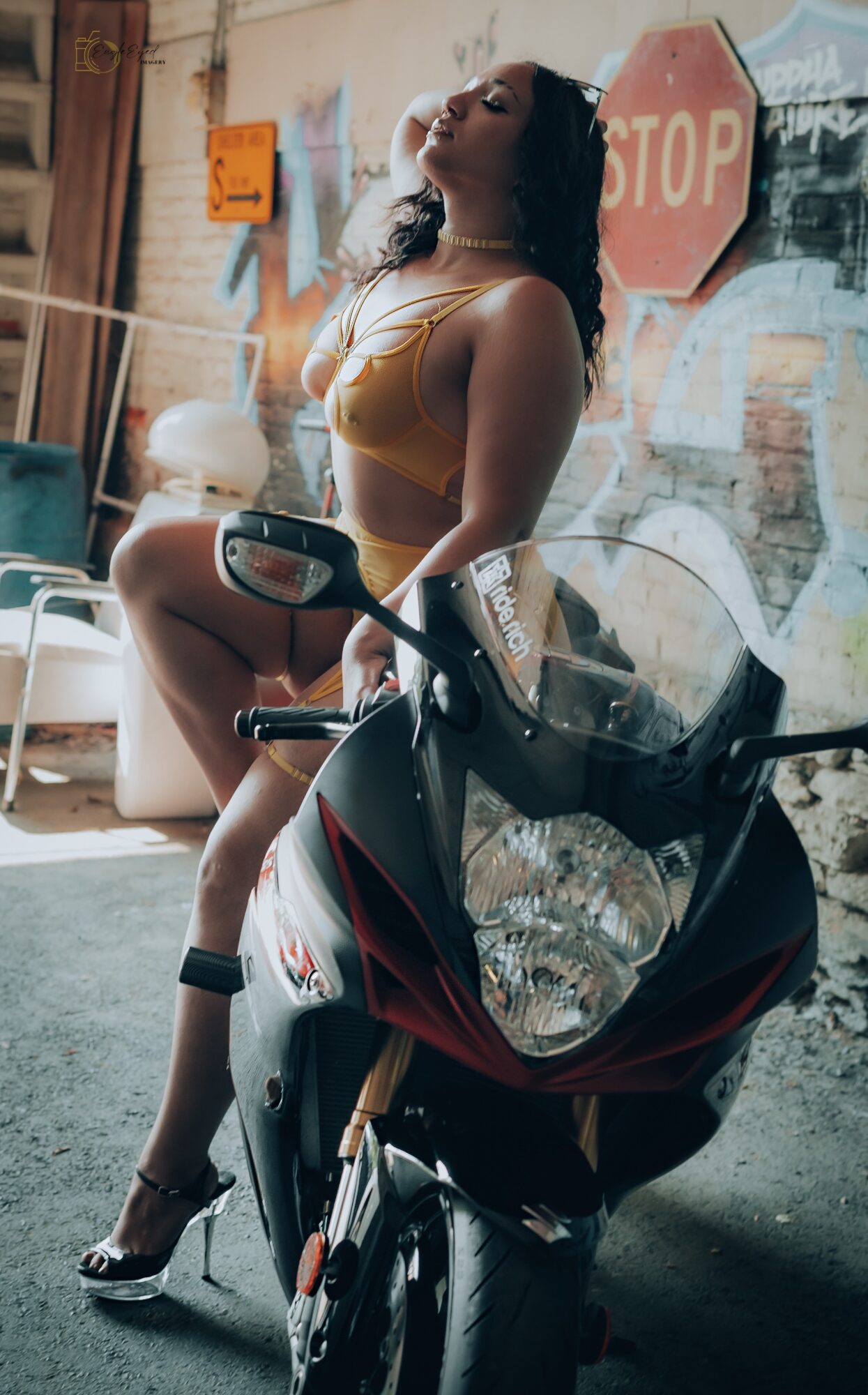 Woman in yellow top and shorts leaning on a motorcycle in an urban setting with a stop sign behind her.