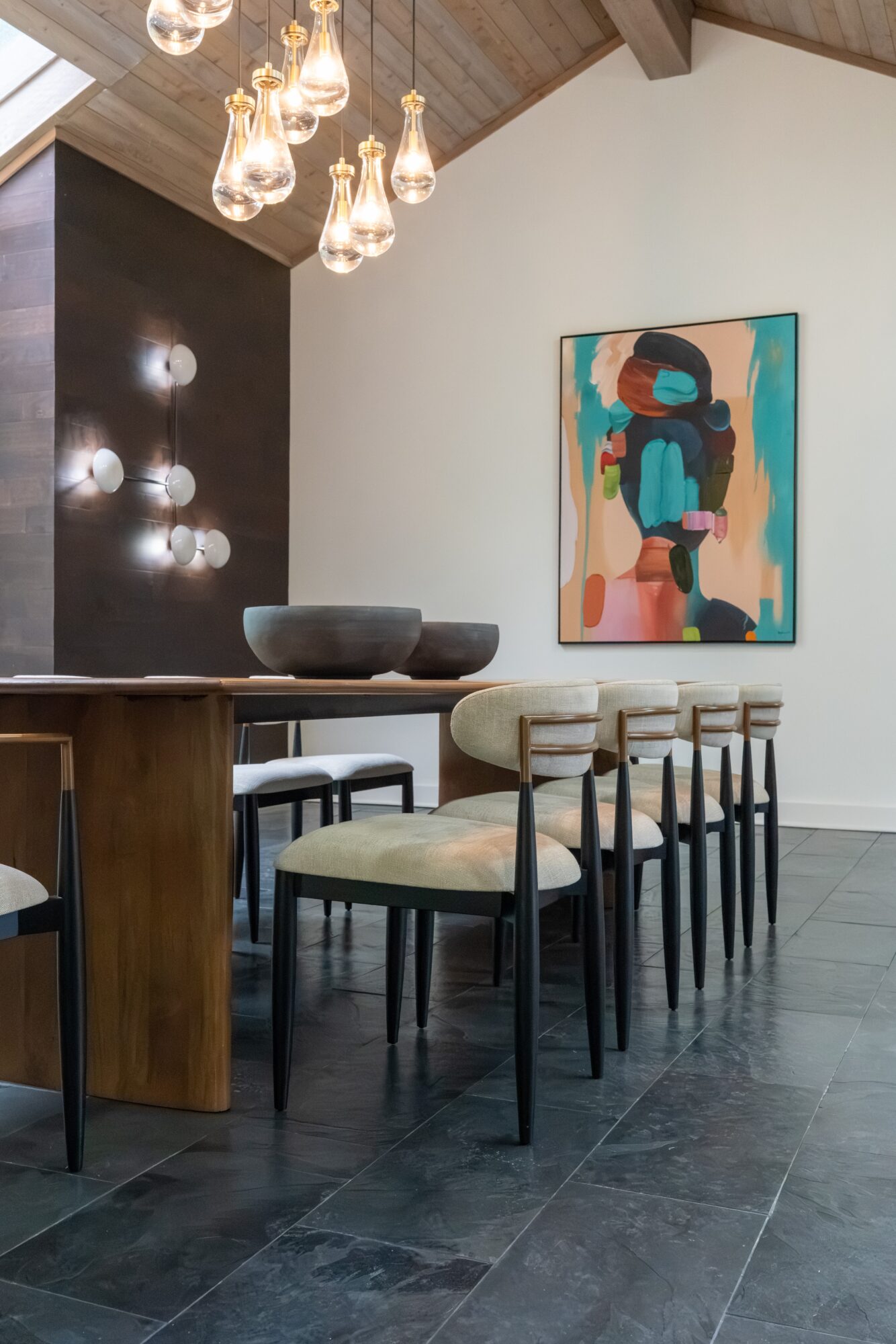 Interior with hanging light bulbs, a black and wood cabinet, a bowl, and a row of chairs with cream cushions, colorful artwork on wall.