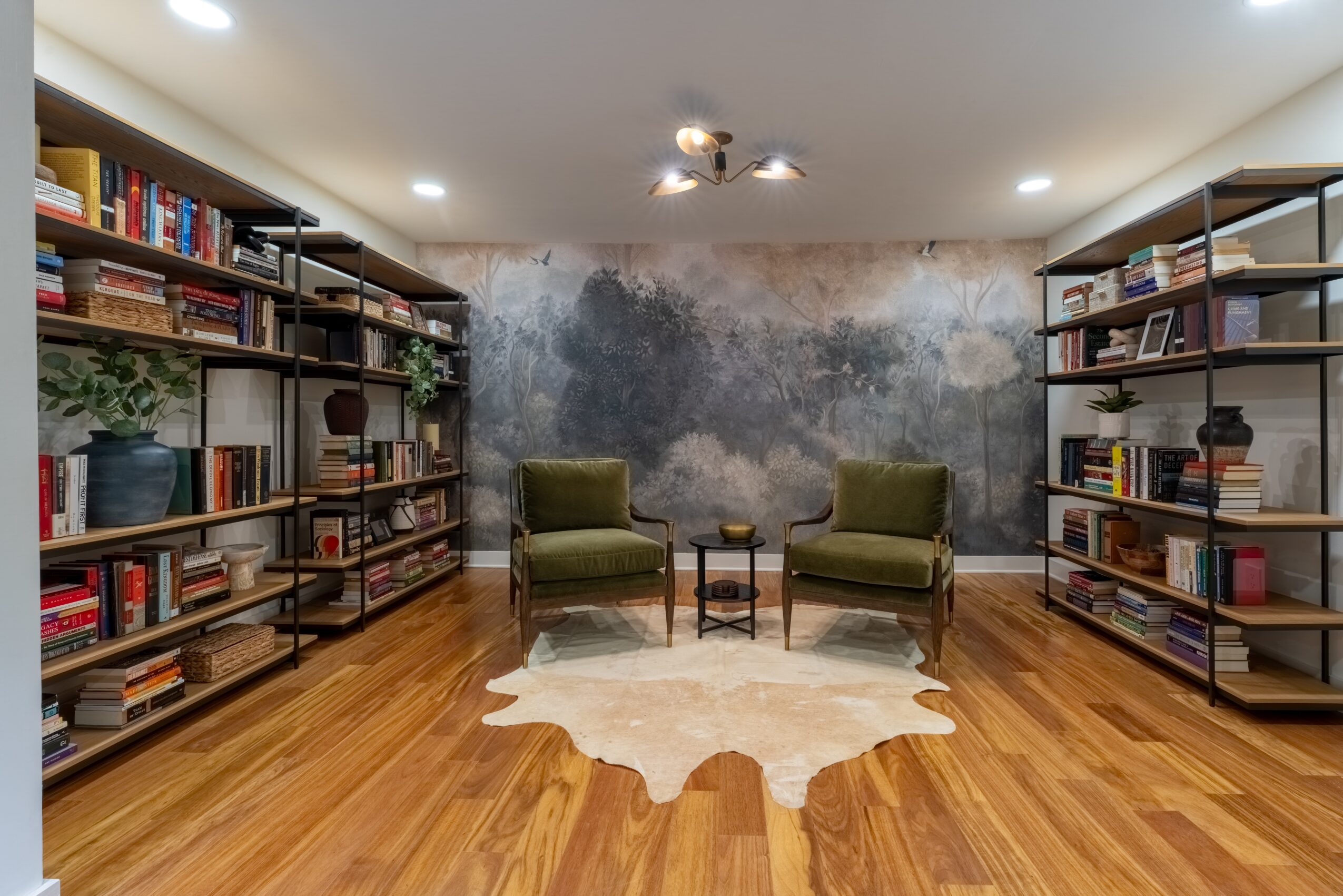 Room with two green armchairs, a small table, bookshelves on both sides, a cowhide rug, and a textured wall mural.