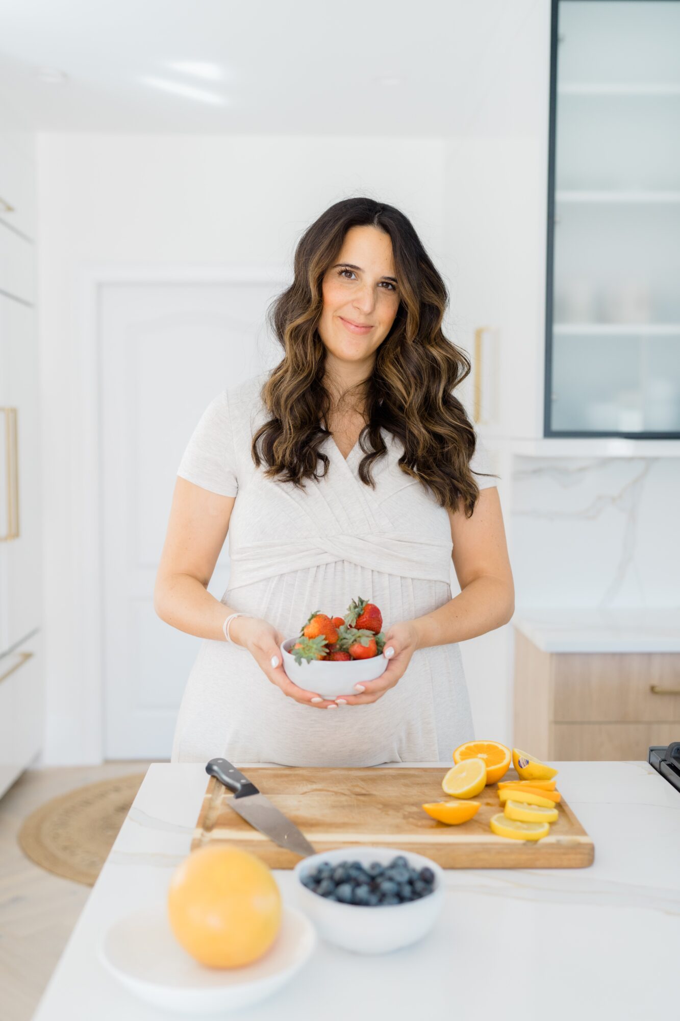Woman with long wavy hair holding a bowl of strawberries in a bright kitchen.