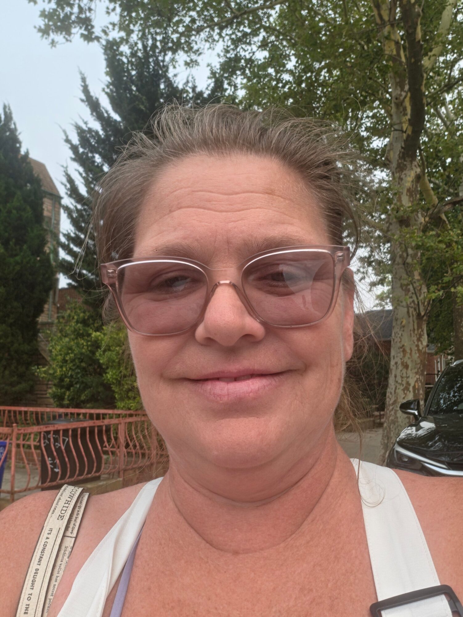 Woman with glasses outdoors, trees and a car in the background, smiling slightly.