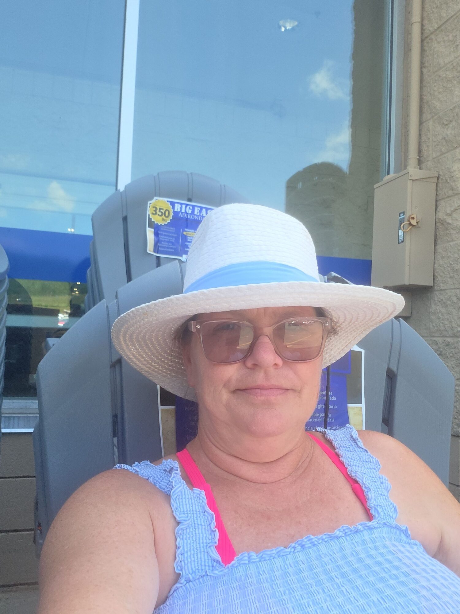 Woman wearing a wide-brimmed hat and sunglasses, sitting outdoors near a glass building, with a blue sky reflected in the window.