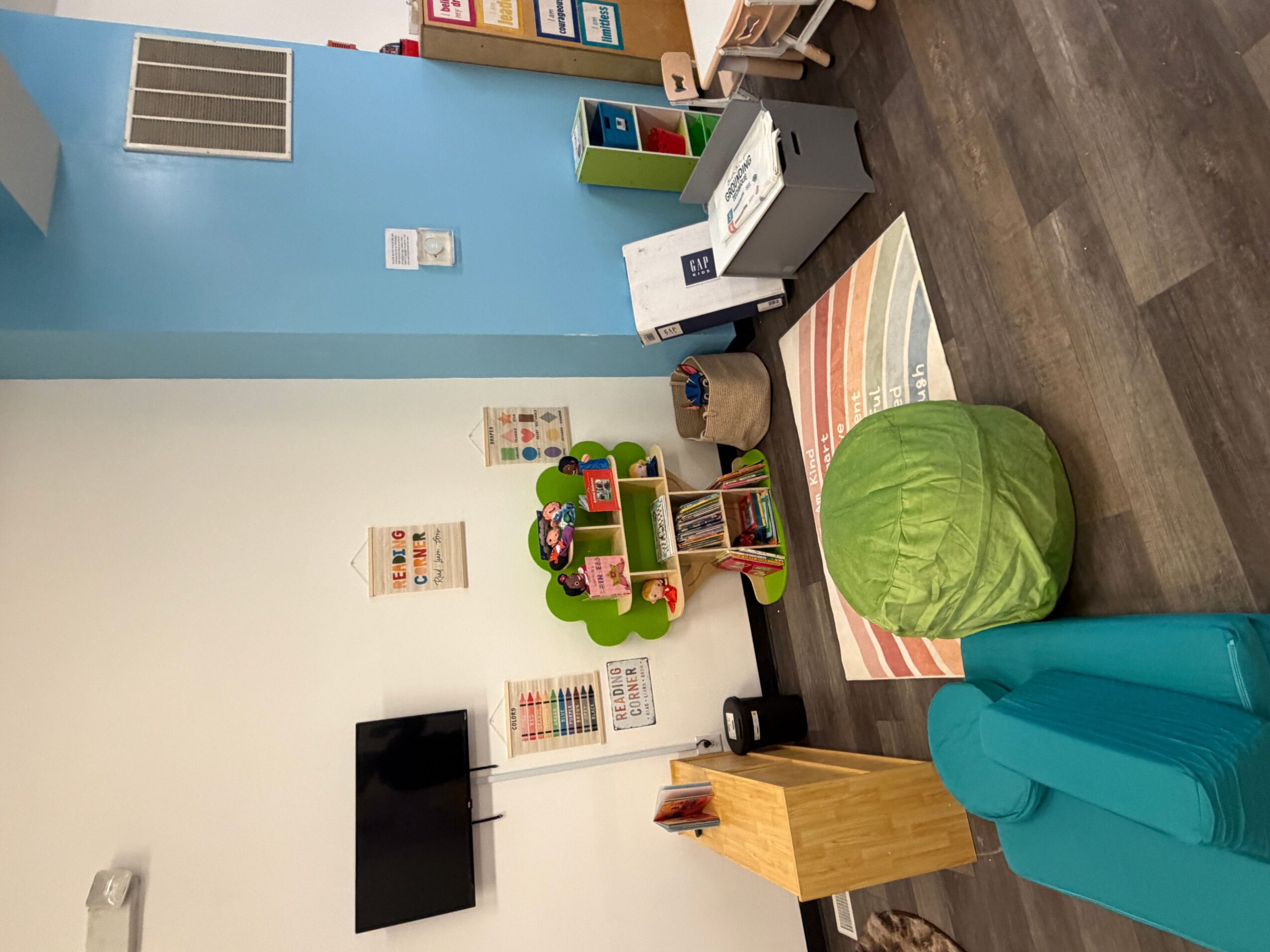 Children's playroom with a green bean bag, teal armchair, small table, and colorful toys on shelves.